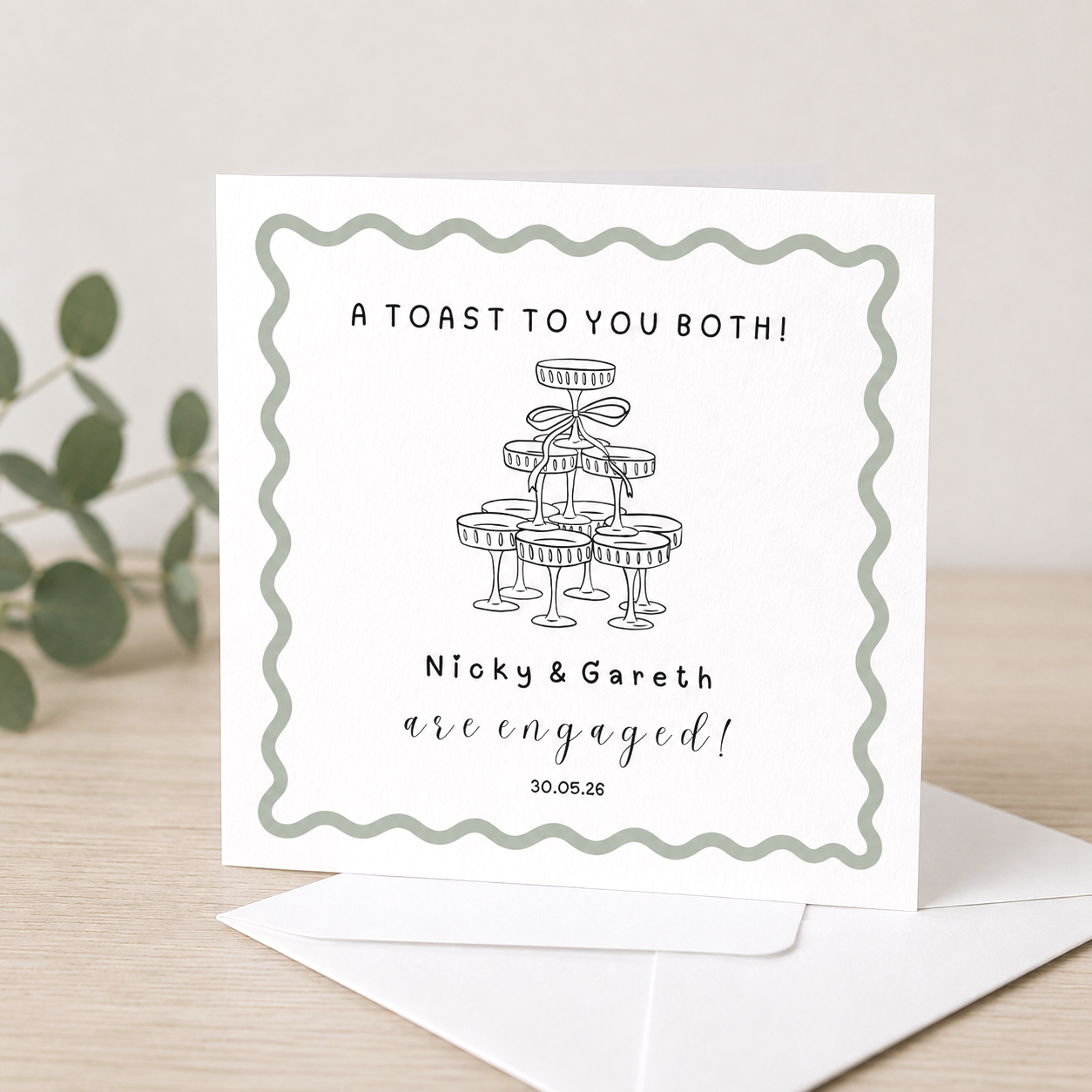 Wedding or engagement announcement card with a champagne glass illustration and text saying 'A toast to you both! Nicky & Gareth are engaged! 30.05.26' on a light-colored table with a white envelope and a green plant in the background.
