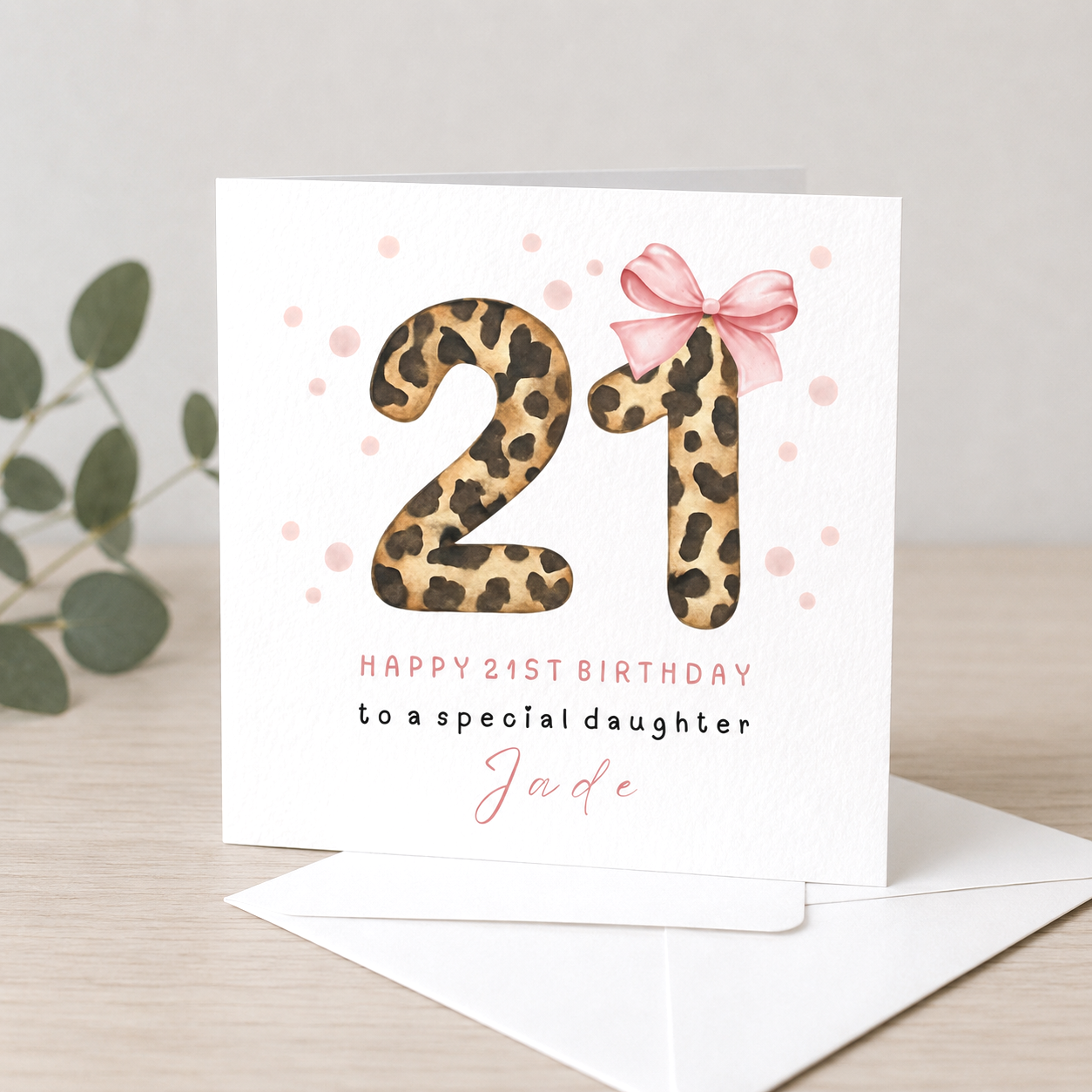 Leopard print number 21 on a birthday card with pink polka dots, pink bow, and text saying 'Happy 21st Birthday to a special daughter Jade' inside a white envelope on a light wooden surface with green leaves nearby.