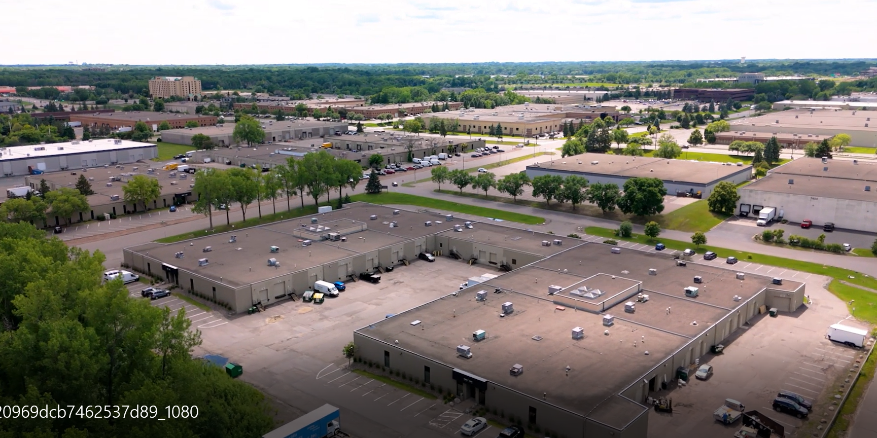 Aerial view of a large industrial or commercial area with multiple warehouses, parking lots, and trees under a partly cloudy sky.