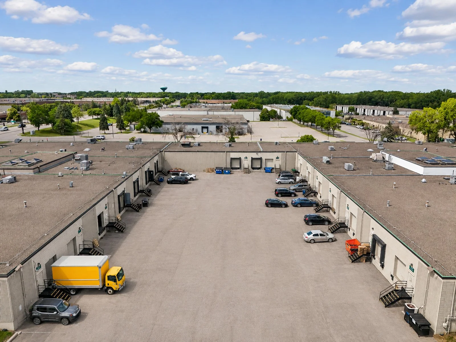An aerial view of a commercial parking lot surrounded by buildings, with several parked cars, loading docks, trees, and a blue sky with clouds in the background.