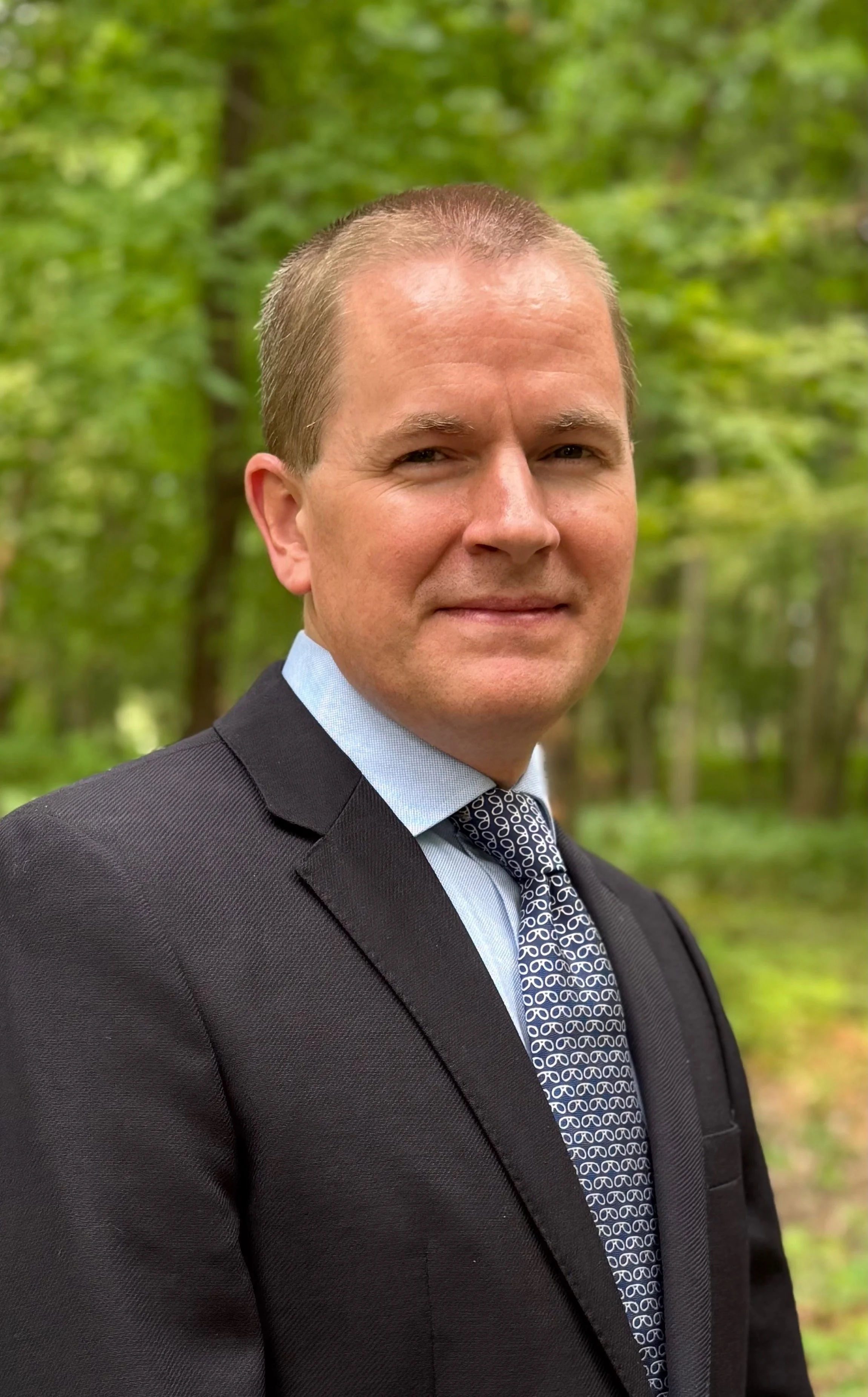 A man wearing a dark suit, light blue shirt, and patterned tie standing outdoors in front of a lush, green forest.