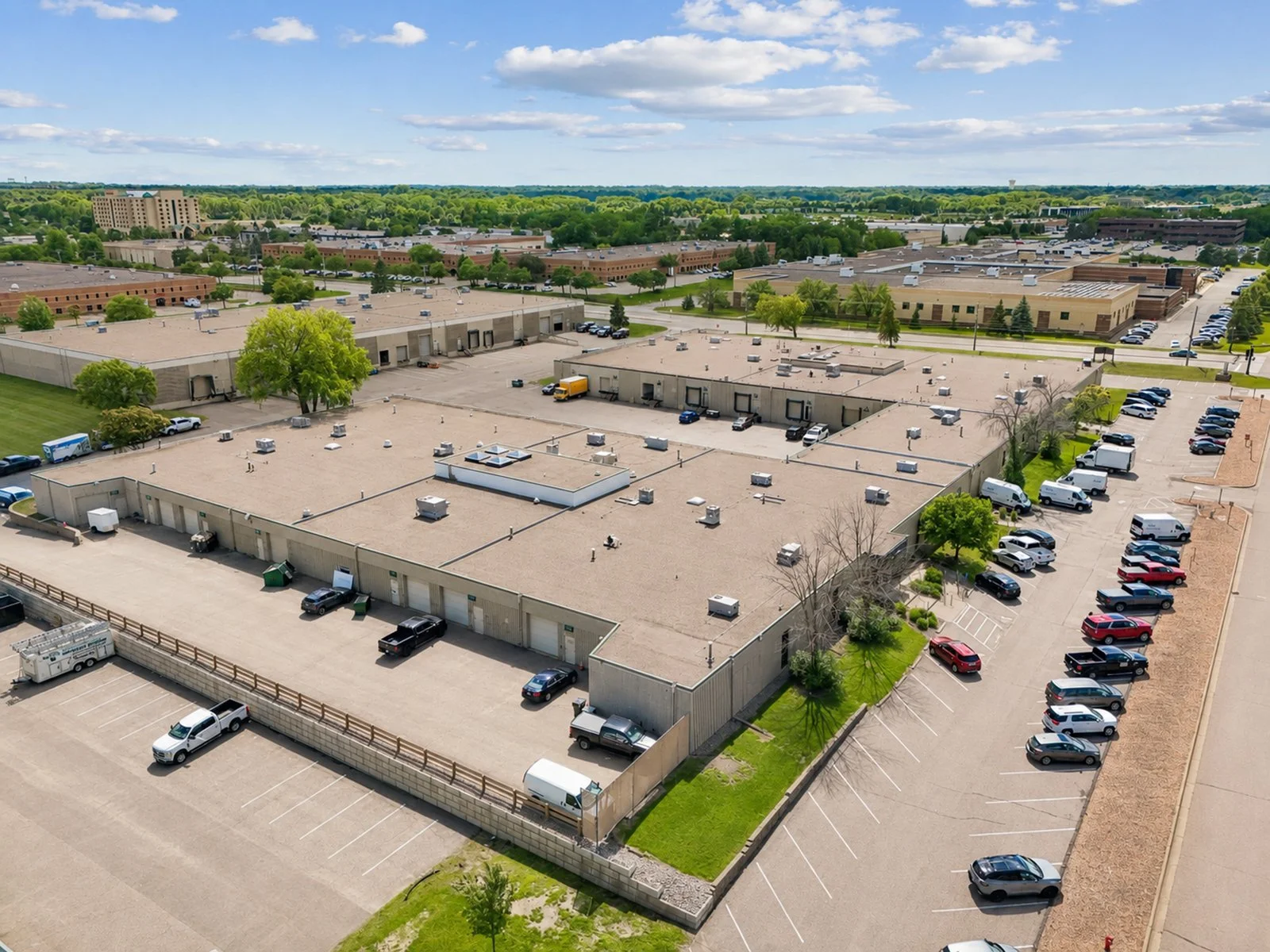 Aerial view of a commercial area with large flat roofs, parking lots, trees, and surrounding buildings under a partly cloudy sky.