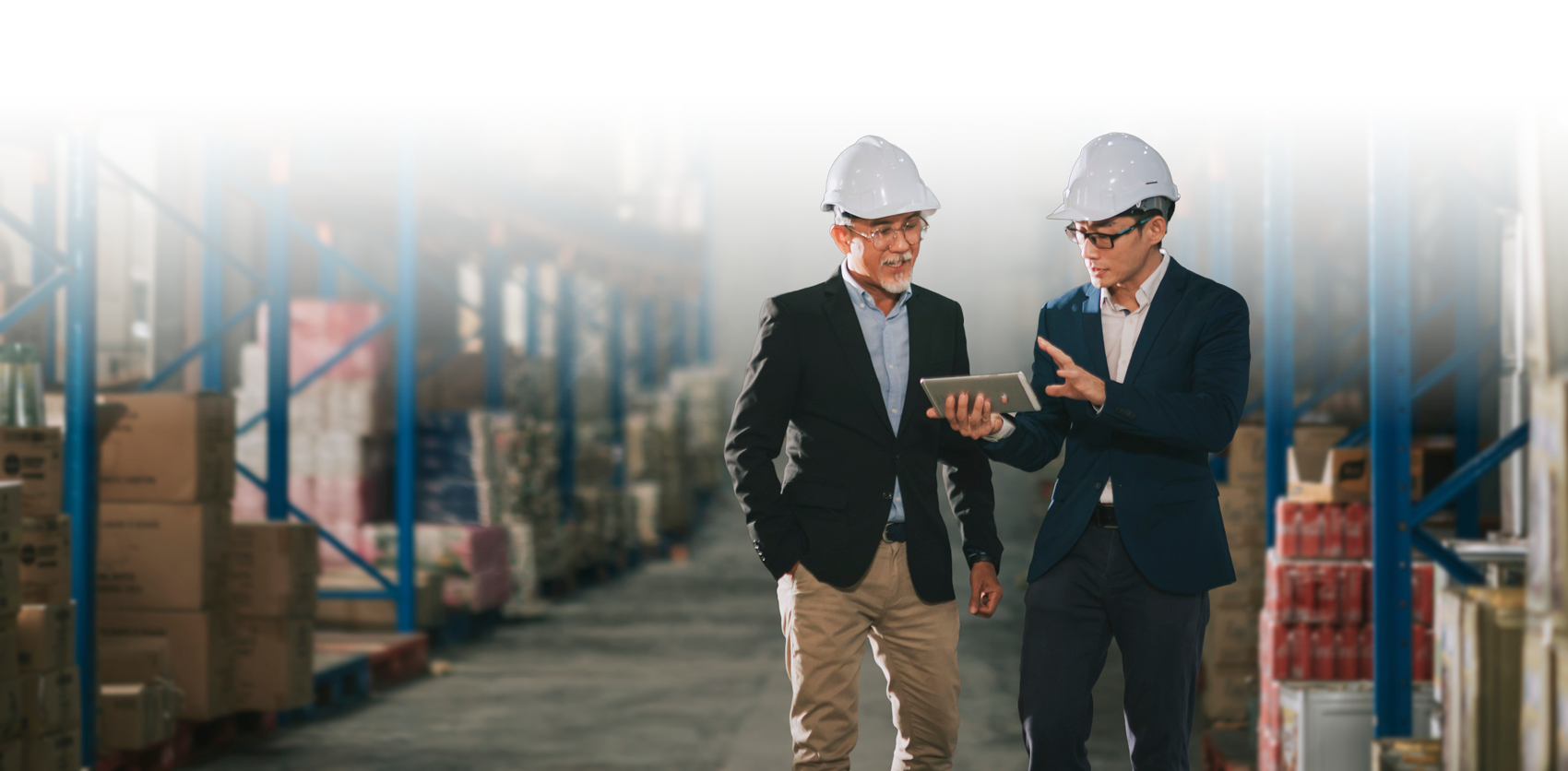 Two men wearing business suits and safety helmets in a warehouse, looking at a tablet together.
