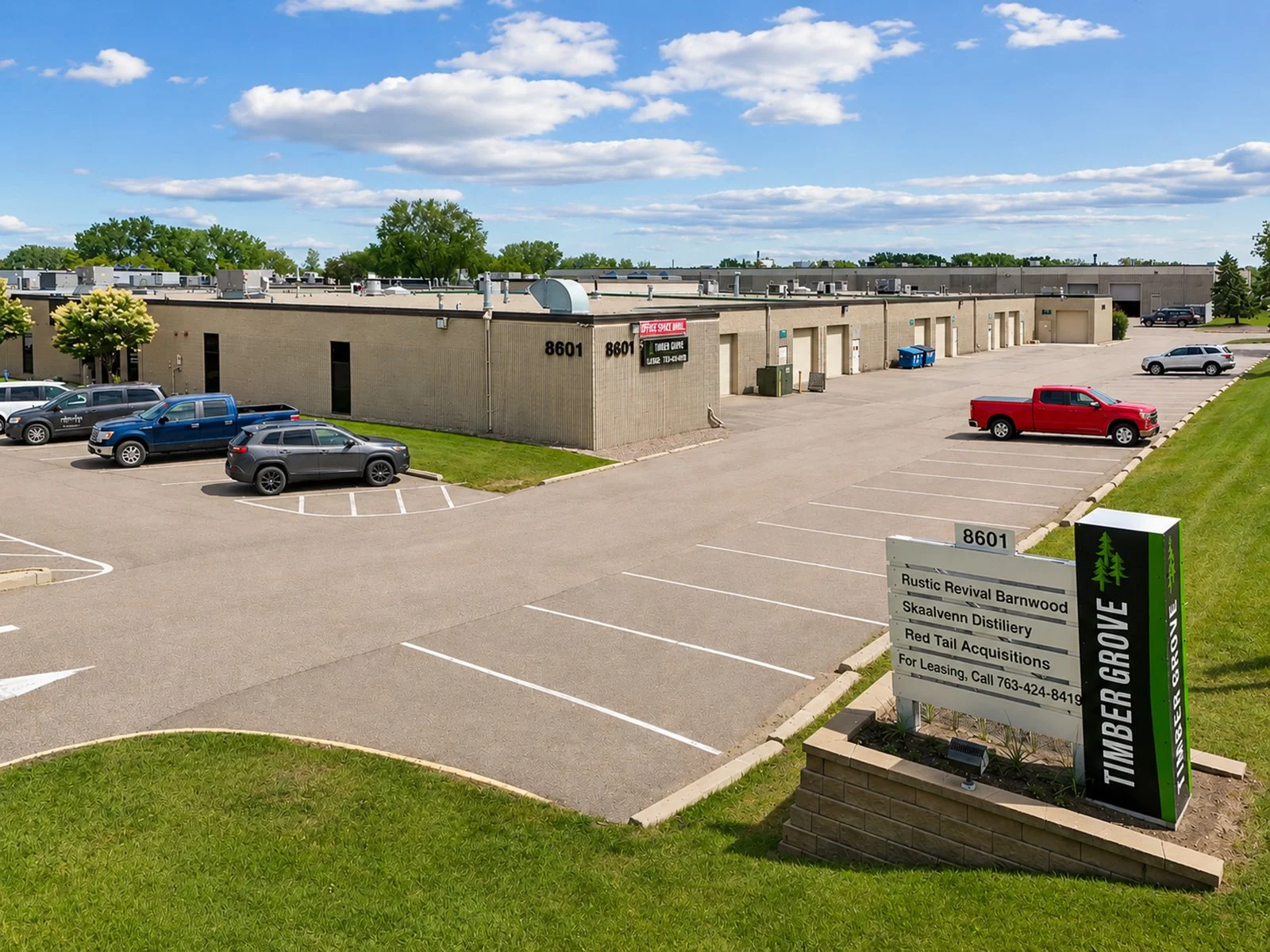 Parking lot outside a business complex with several parked cars and a building in the background. A sign displays the names of businesses including Rustic Revival Barnwood and SkaaLavenn Distillery.