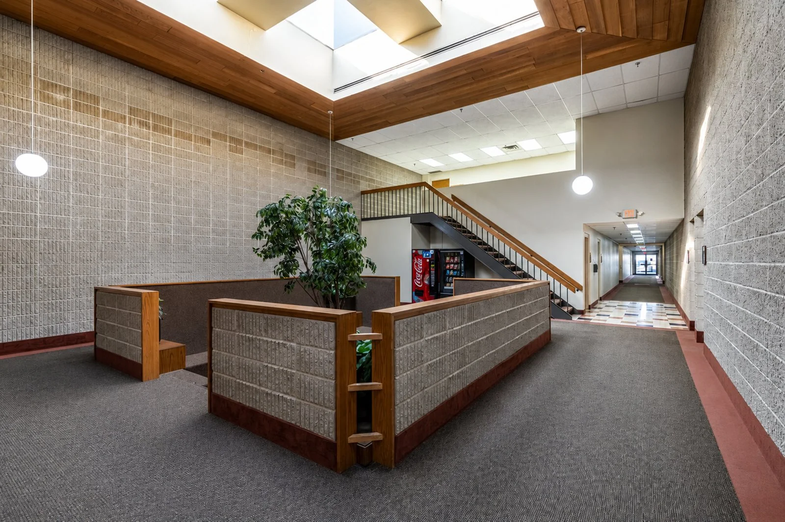 An indoor hallway with beige brick walls, a wooden ceiling, and a skylight. There's a staircase with wooden handrails, a vending machine, a potted plant, and hanging round light fixtures. A corridor extends into the distance.