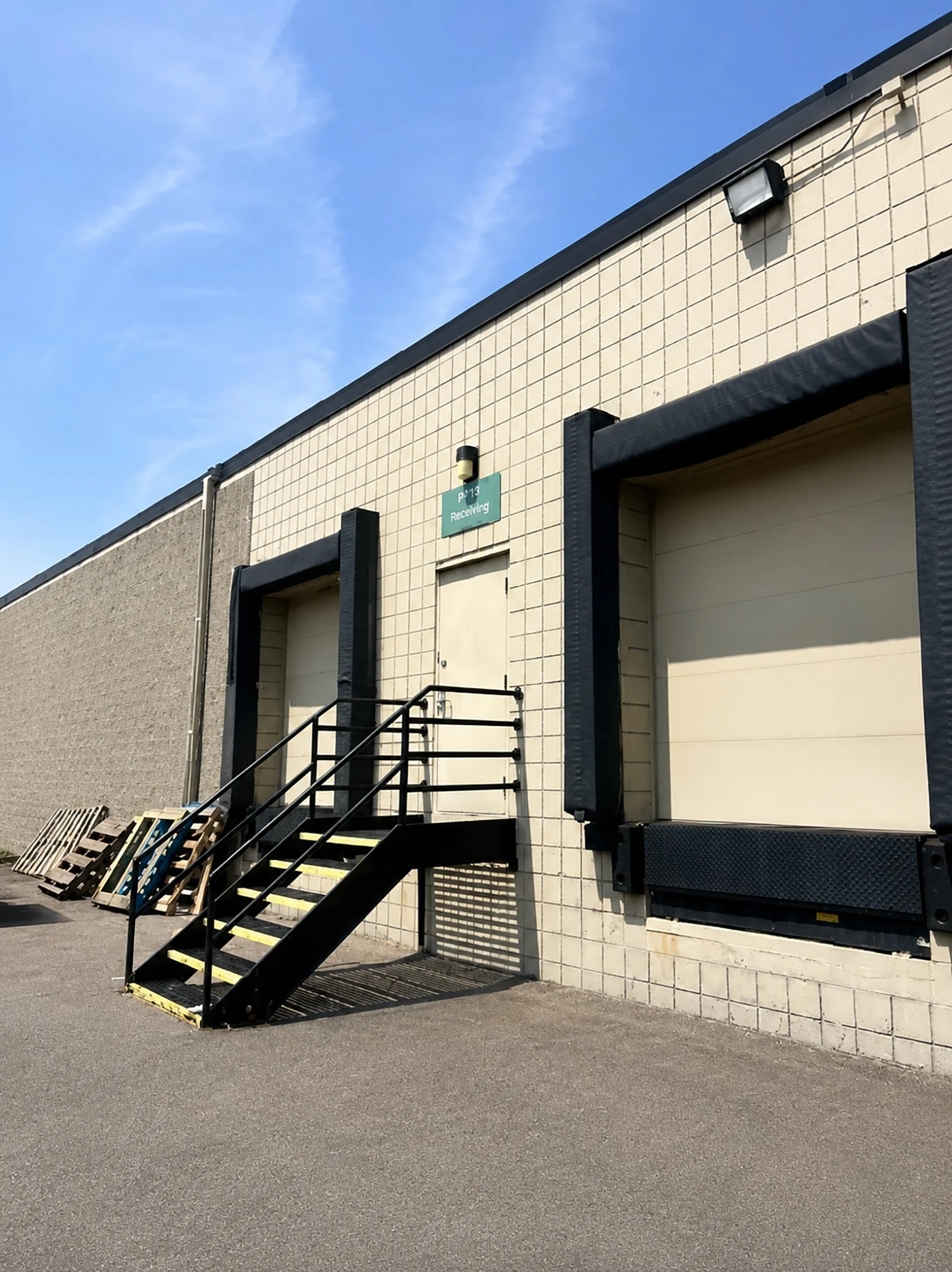 Loading dock with two garages, black stairs with yellow safety markings, pallets, and a beige brick building under a clear blue sky.