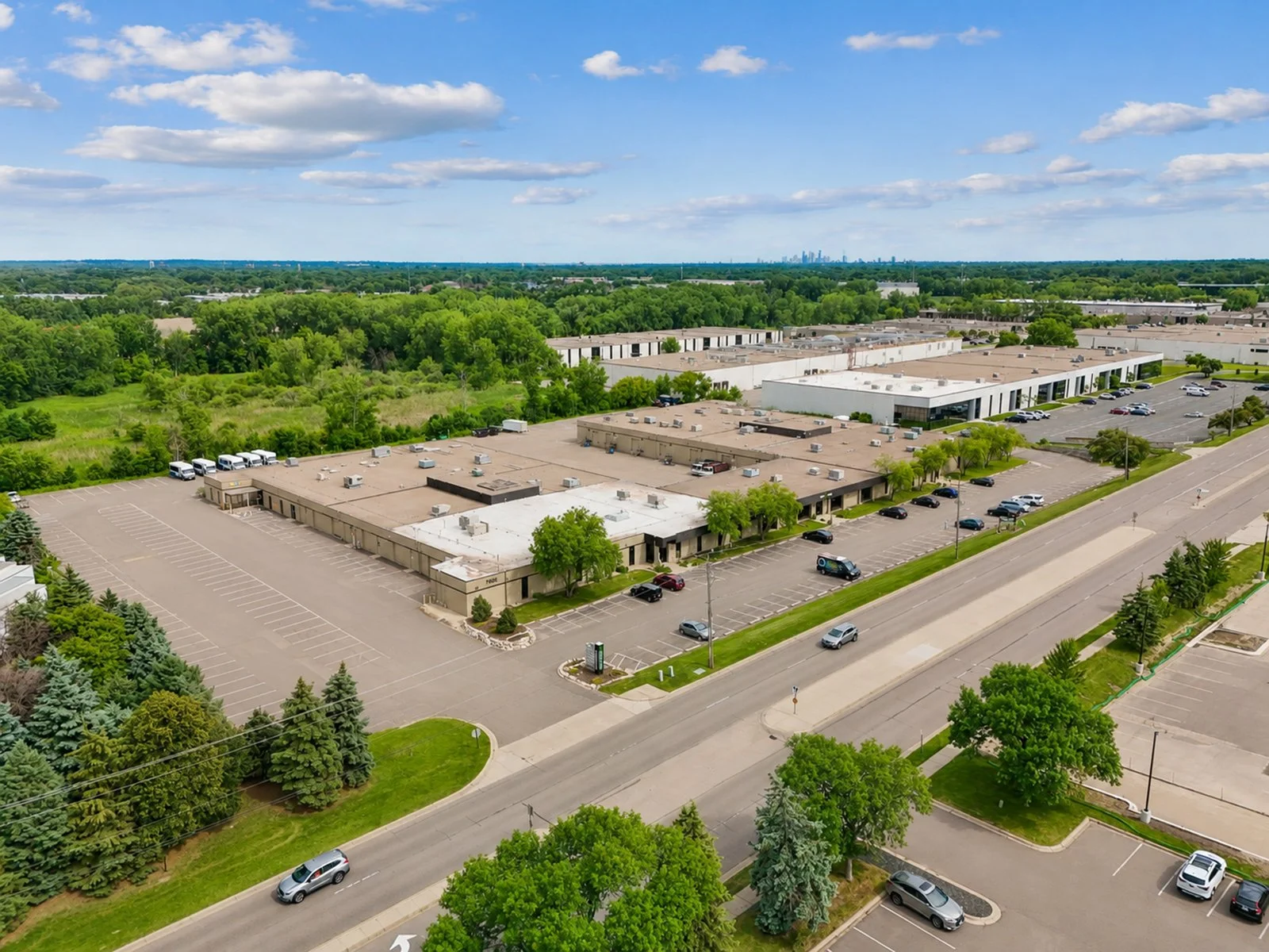 An aerial view of commercial buildings with parking lots, green trees, and a lush forest in the background, under a partly cloudy sky.