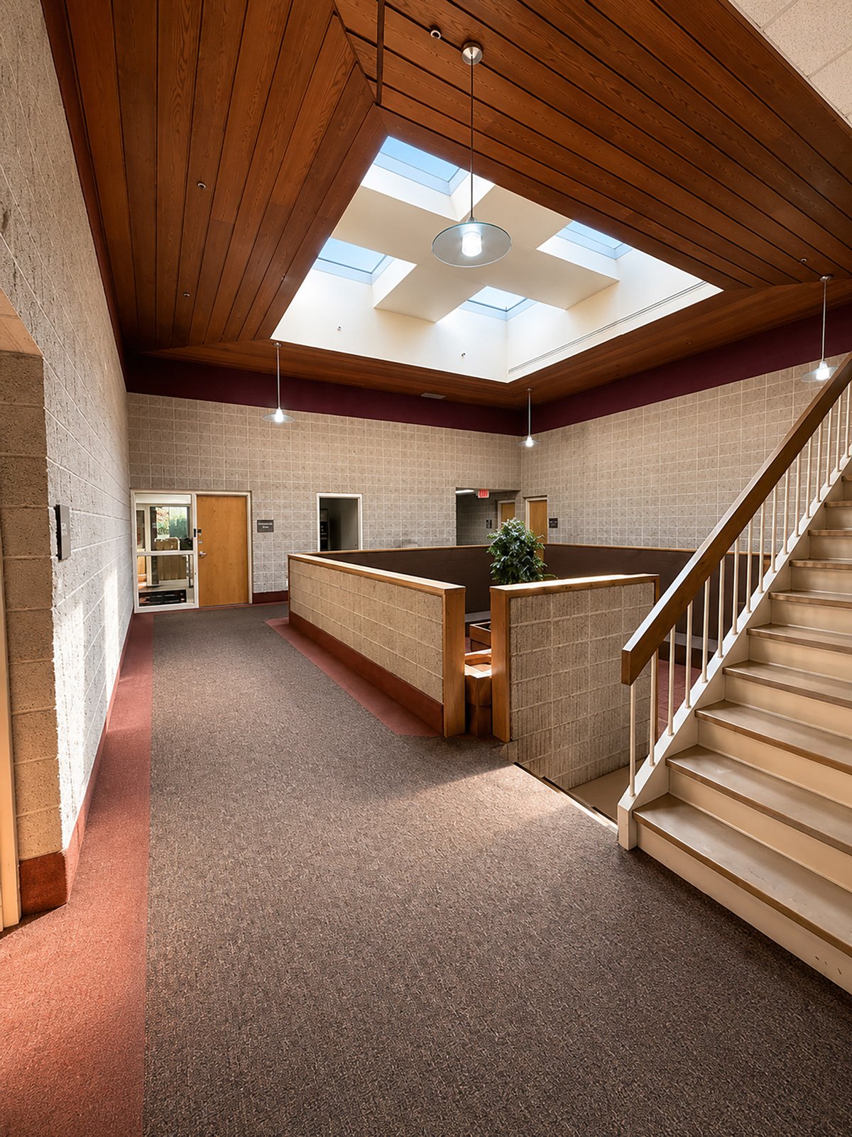 Interior view of a building with a skylight ceiling, carpeted floor, staircase, and walls with beige brick pattern. There are hanging lights and a potted plant near the staircase.