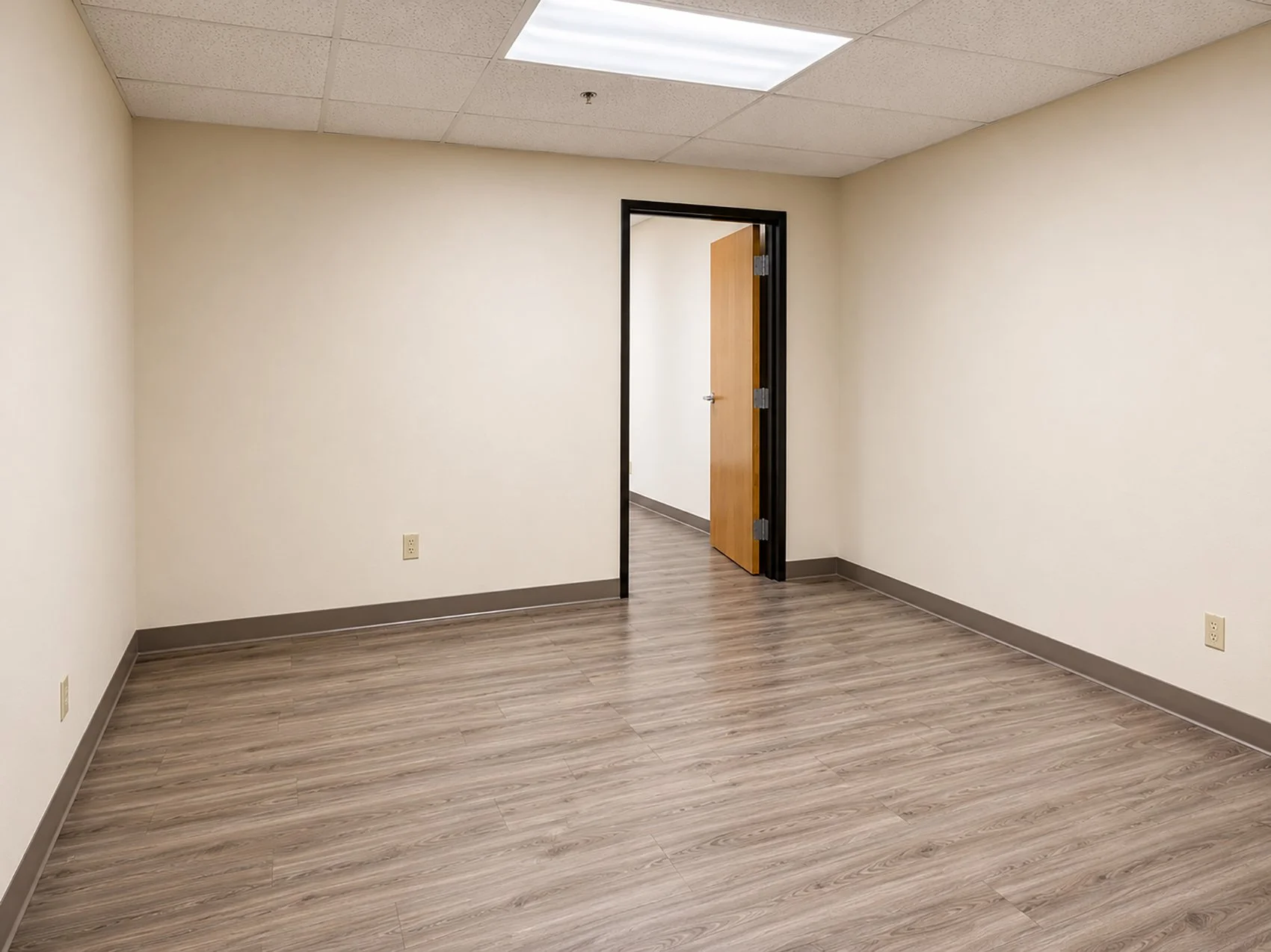 Empty office room with beige walls, wood flooring, and an open door leading to a hallway.