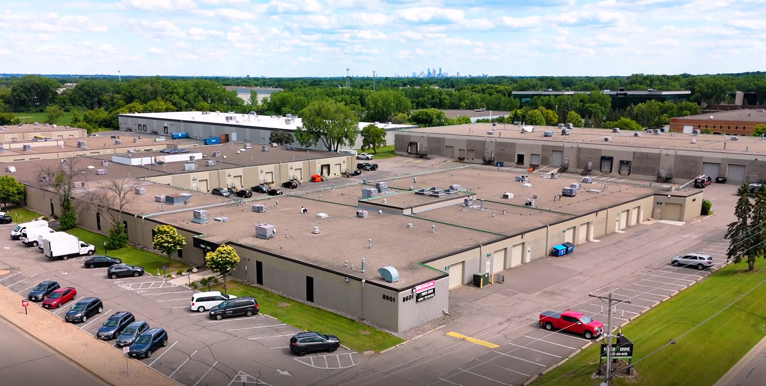 An aerial view of a large commercial building with a parking lot, surrounded by greenery, with a city skyline visible in the distance under a partly cloudy sky.