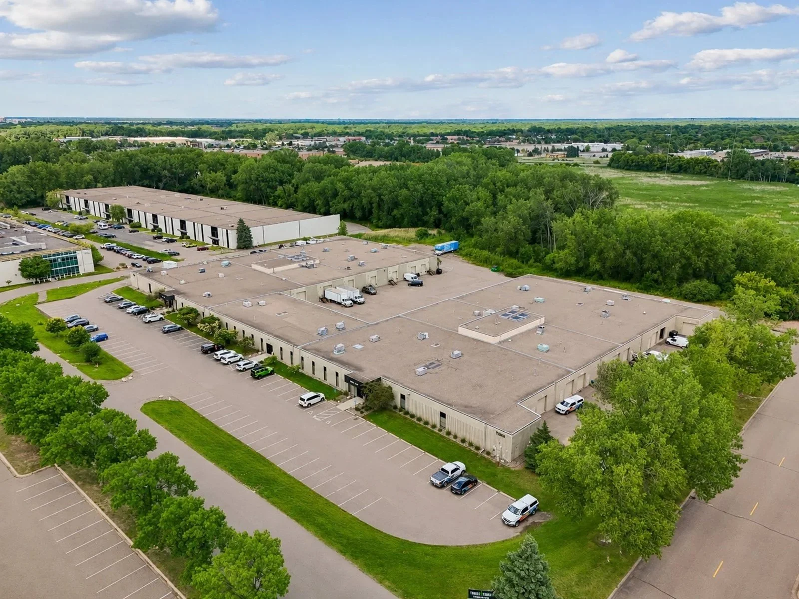 Aerial view of a large commercial building with a flat roof and parking lots, surrounded by green trees and open space.