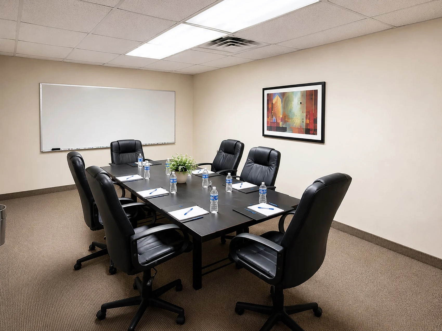 Conference room with a long dark wooden table, eight black leather chairs, water bottles, notepads, pens, a potted plant in the center, a whiteboard on the wall, a framed colorful abstract painting, beige walls, and a ceiling with fluorescent lighting and an air vent.