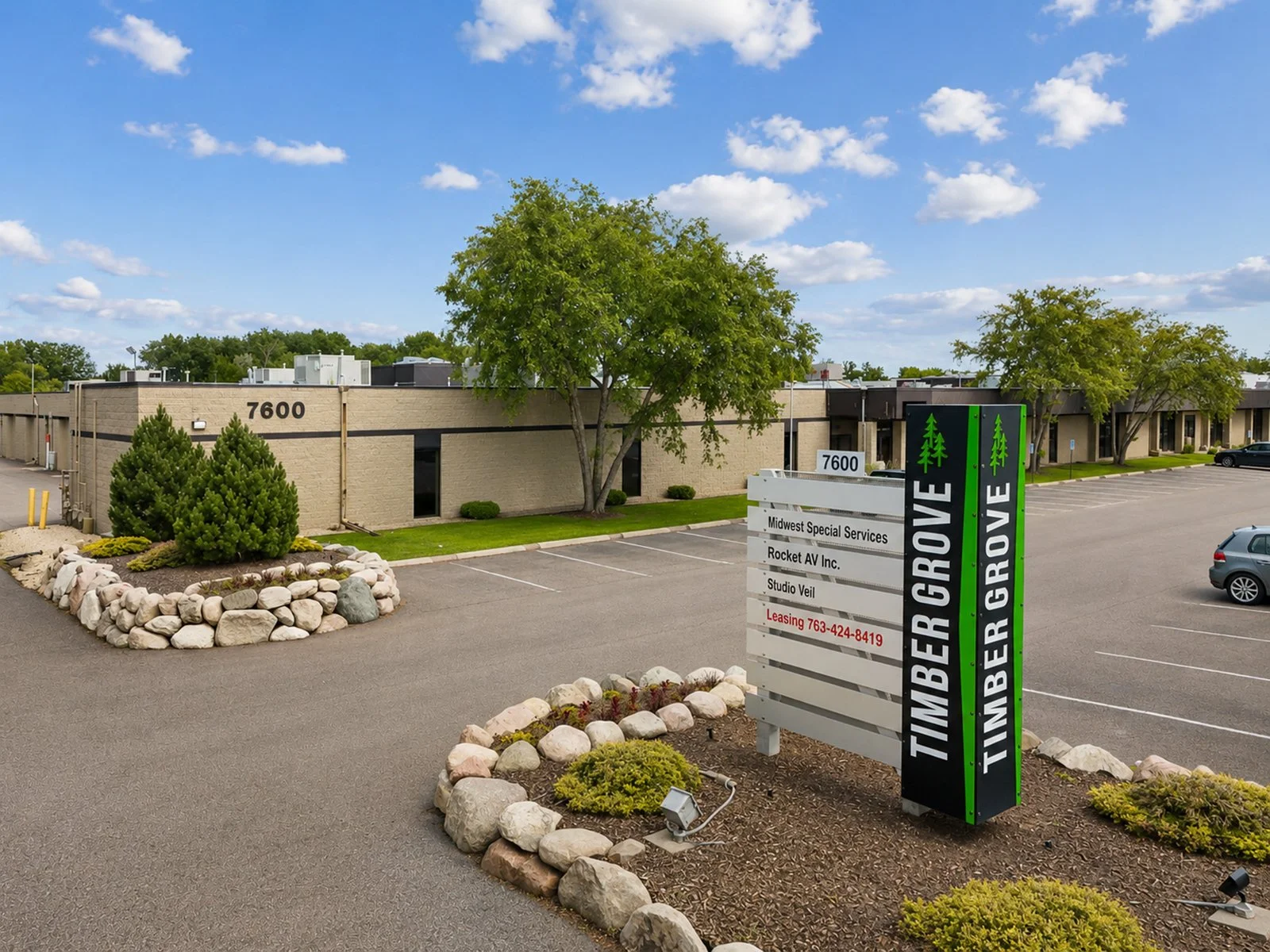 Parking lot of an office building with a sign displaying tenant names, trees, and landscaped rock beds under a partly cloudy sky.