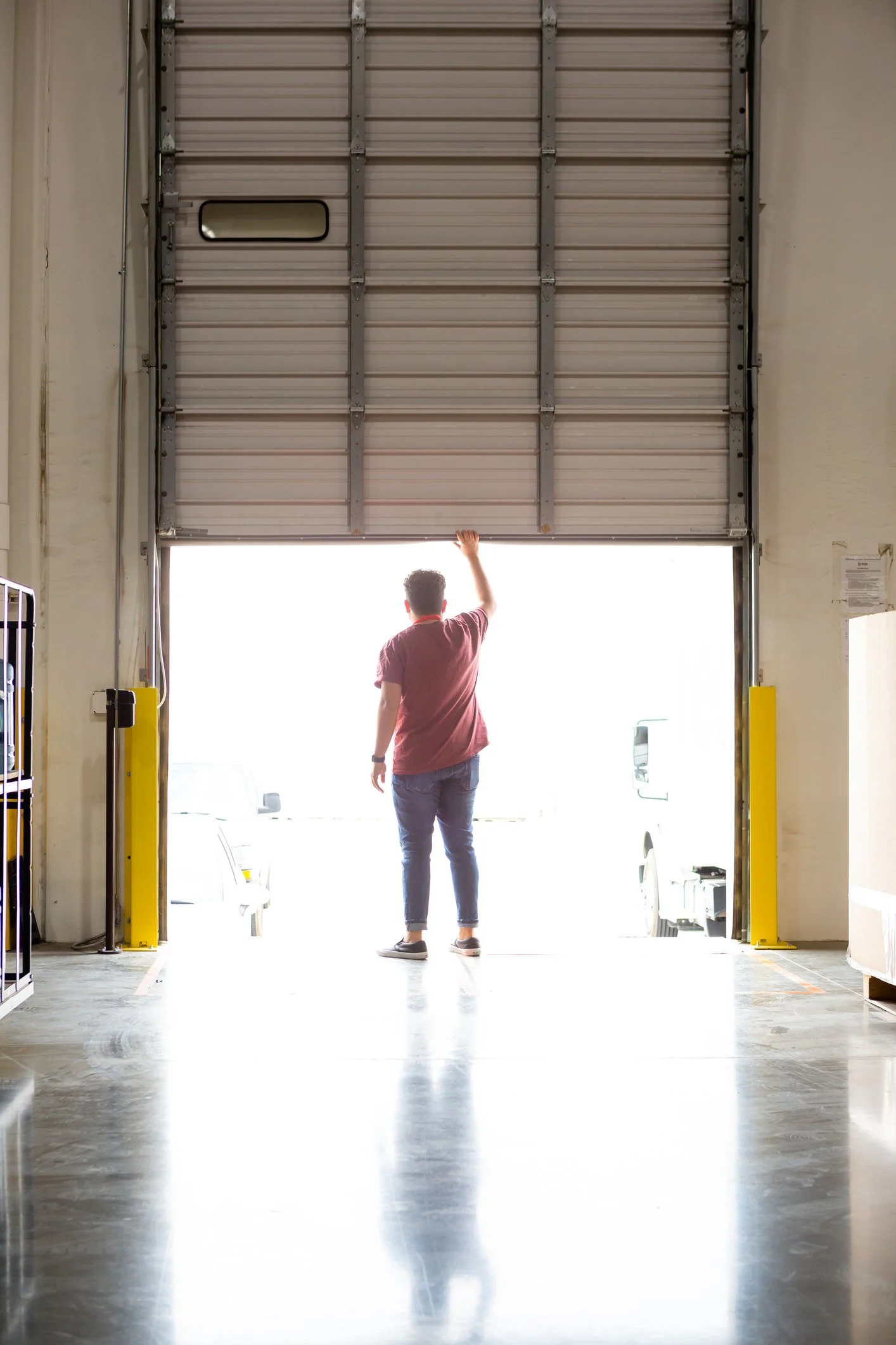 A person in casual clothes is standing in a warehouse near an open garage door, looking outside as sunlight floods the space.