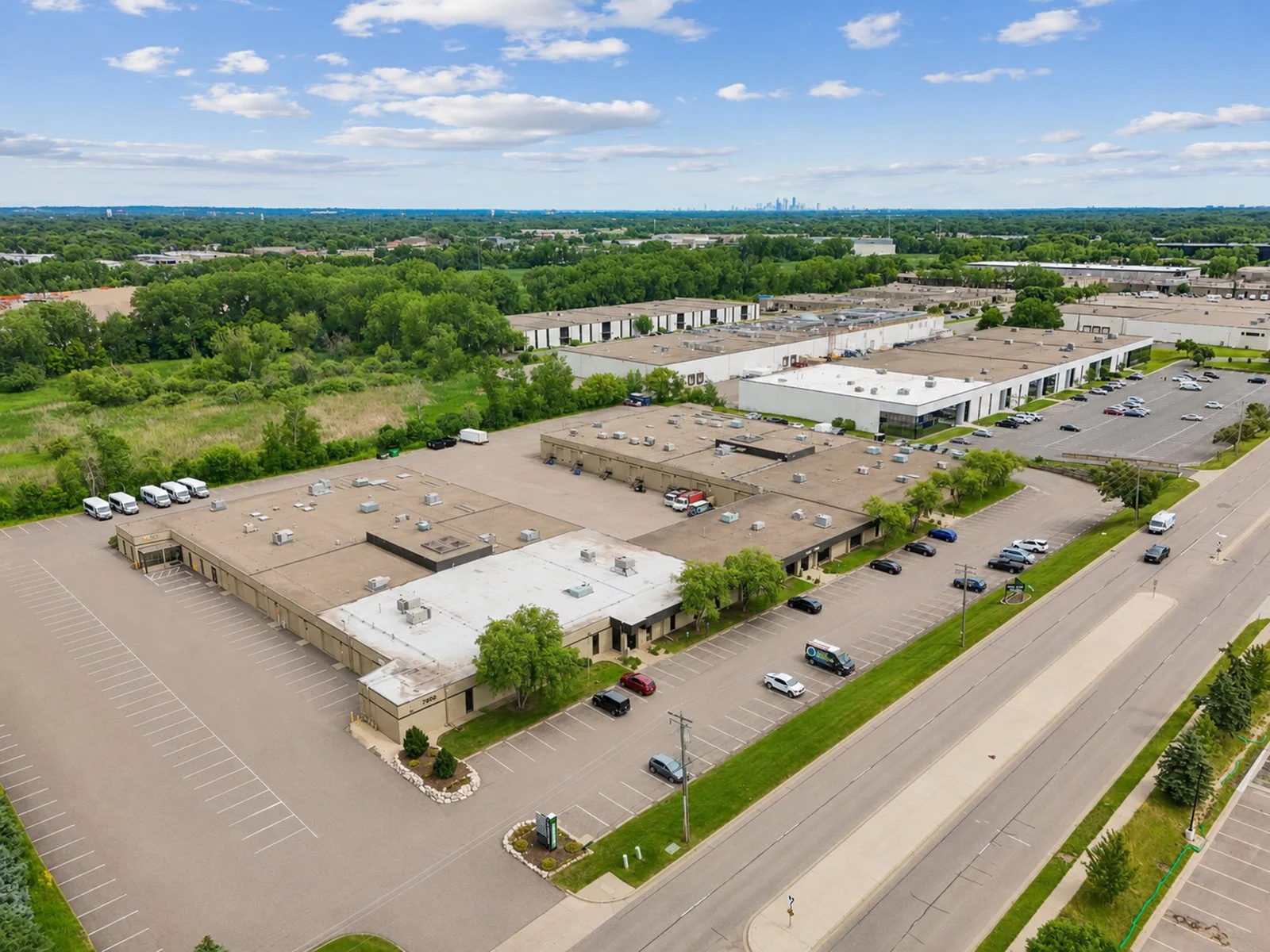 An aerial view of a large commercial building complex with multiple parking lots, surrounded by greenery and trees, under a partly cloudy sky.