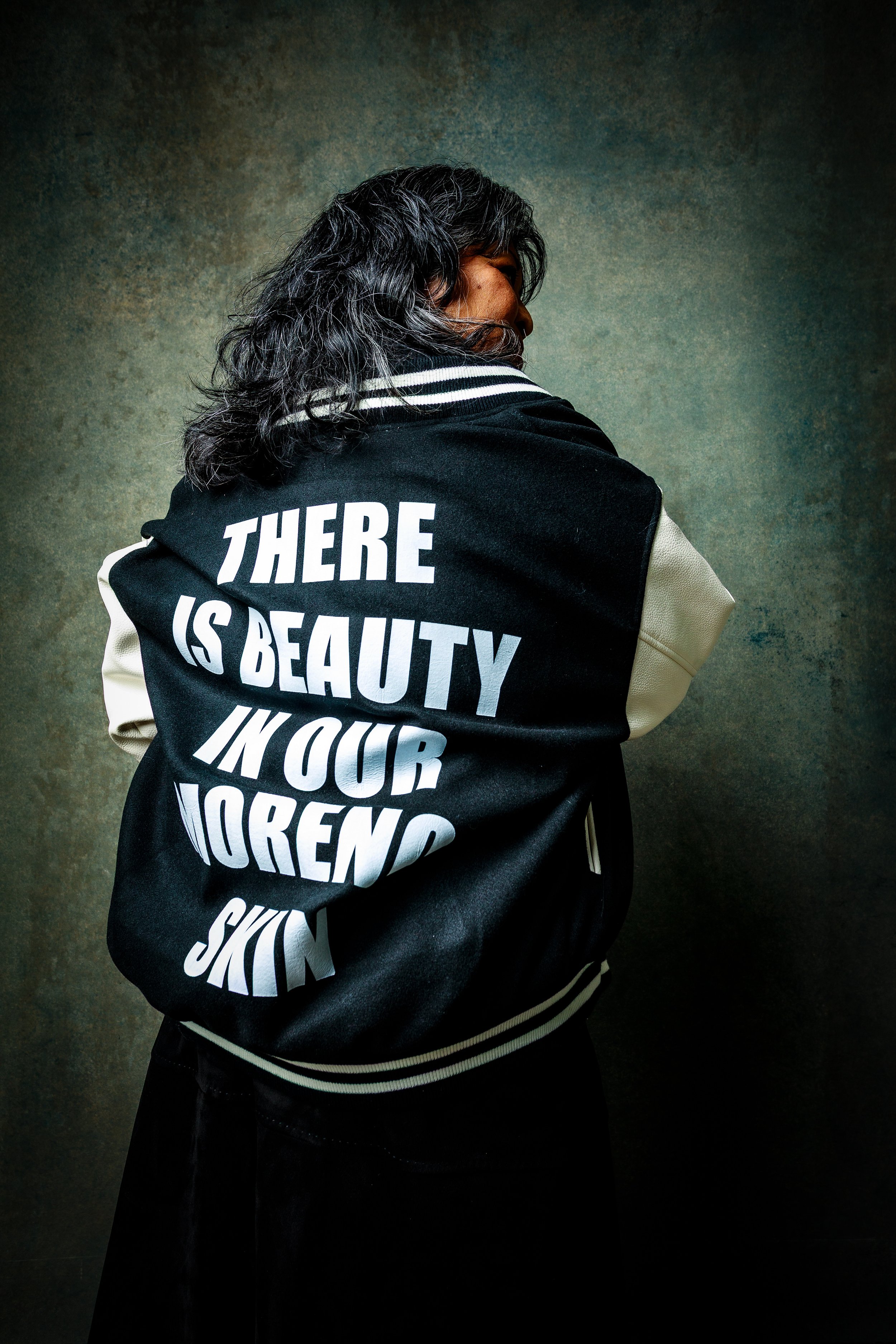 Person with long, curly black hair wearing a black and cream varsity jacket with white text on the back that reads 'THERE IS BEAUTY IN OUR MORENA SKIN' standing against a textured grayish-green background.