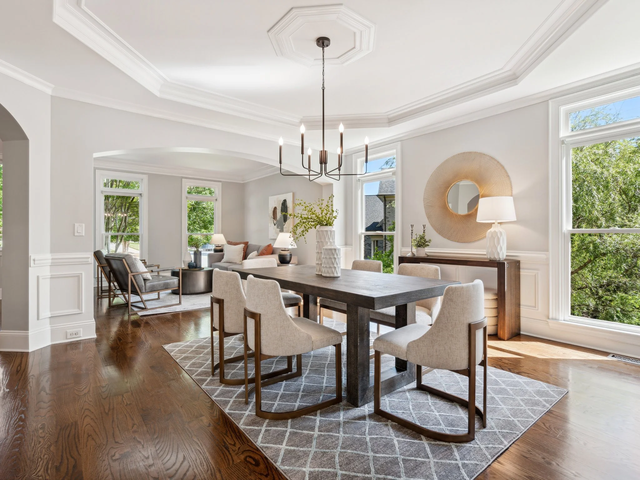 A spacious, well-lit dining room and living area with large windows showing green trees outside. The room features a dark wood dining table surrounded by beige chairs, modern light fixtures, and decorative elements such as a round mirror, table lamps, and vases.