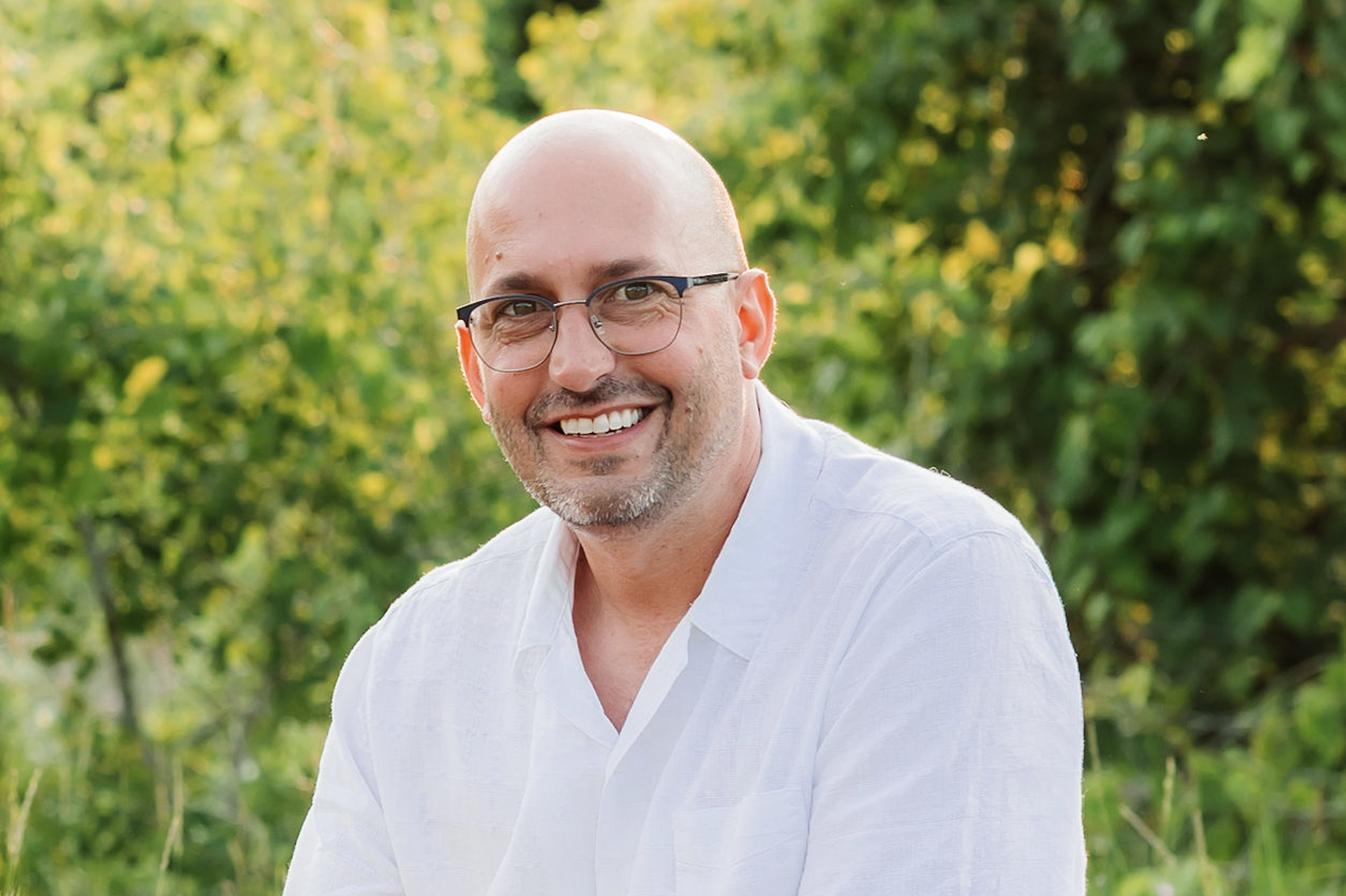 A smiling bald man with glasses wearing a white shirt; greenery background.