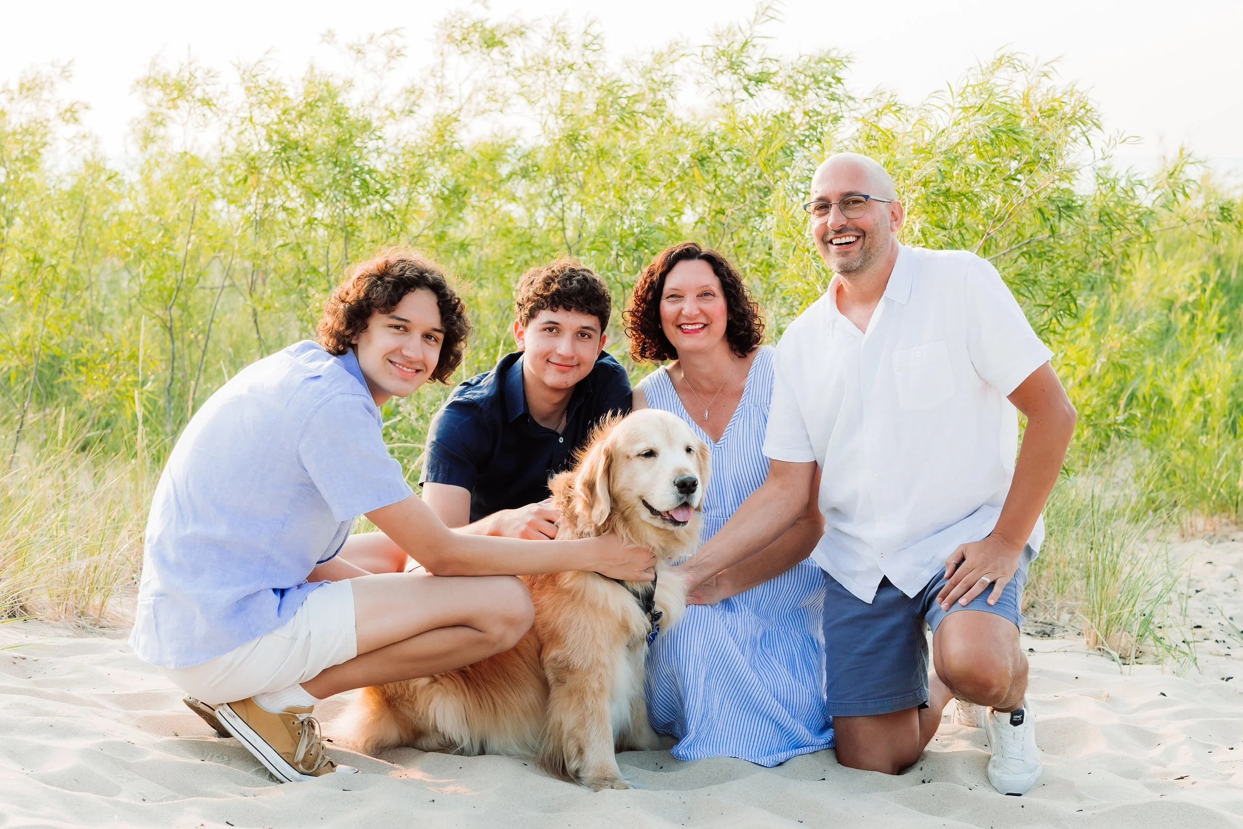 A family with two teenagers, a woman, a man, and a golden retriever dog on a sandy beach with green shrubbery in the background.