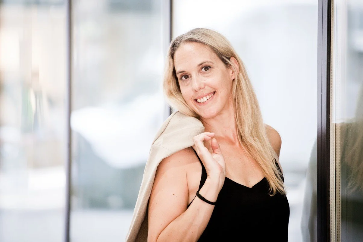 Eine lächelnde Frau mit blonden Haaren, die ein schwarzes Top trägt und eine beige Jacke über die Schulter hält.