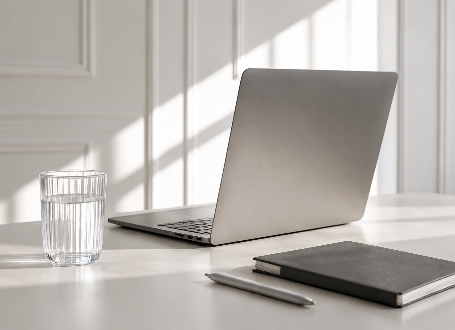 Ein silberner Laptop auf einem weißen Tisch neben einem schwarzen Notizbuch, einem weißen Stift und einem Glas Wasser, vor einem Fenster mit Sonnenlicht.
