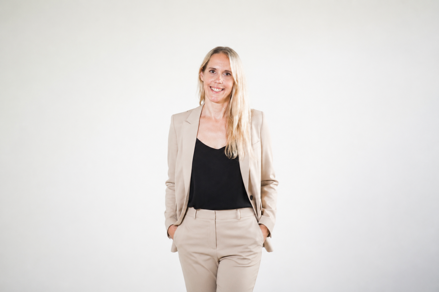 Eine lächelnde Frau in einem beige Business-Anzug mit schwarzen Top steht vor einem weißen Hintergrund.