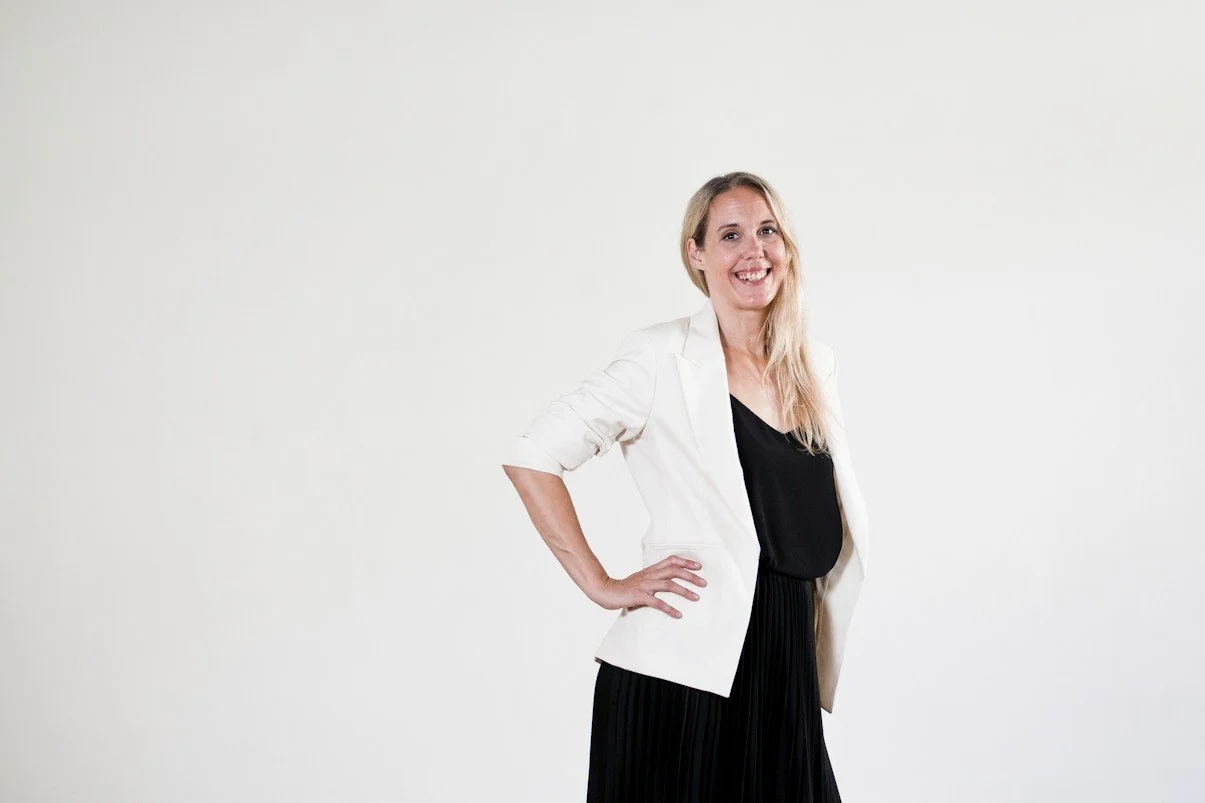 Eine lächelnde Frau in einem weißen Blazer, schwarzem Top und schwarzen Rock steht vor einem weißen Hintergrund.