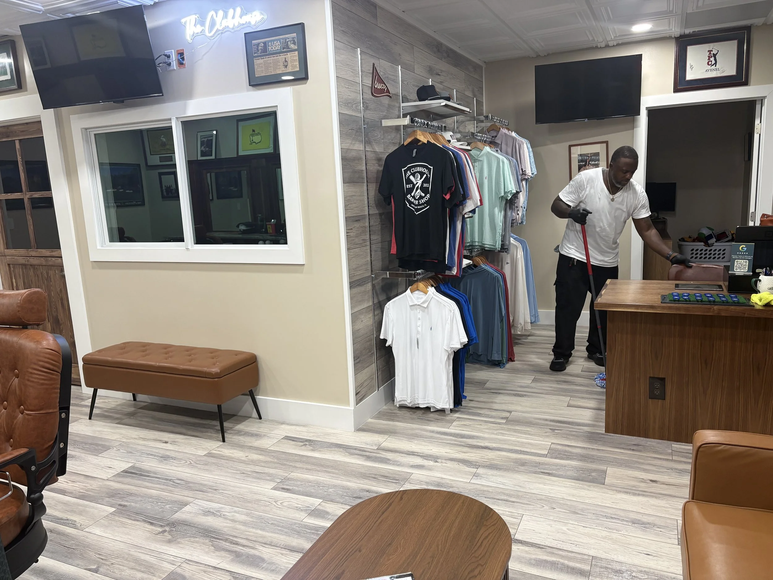 Interior of a clothing store with racks of polo shirts and other apparel, two large flat-screen TVs on the walls, framed posters and signs, a window with a view into another room, and a man cleaning the floor.