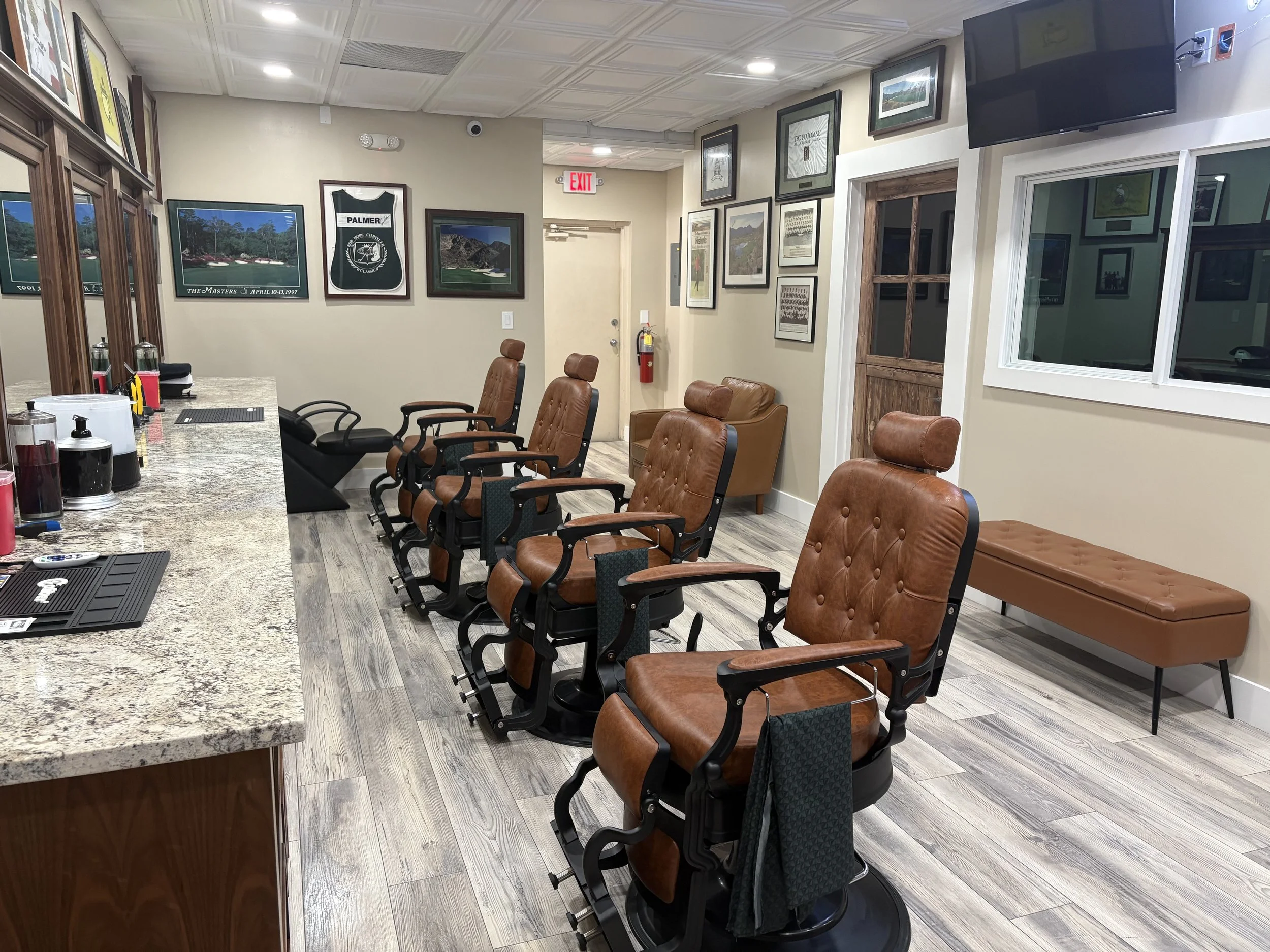 Interior of a clubhouse or lounge area with a bar counter, wood and leather chairs, framed photos and artwork on the walls, a mounted television, and large windows.