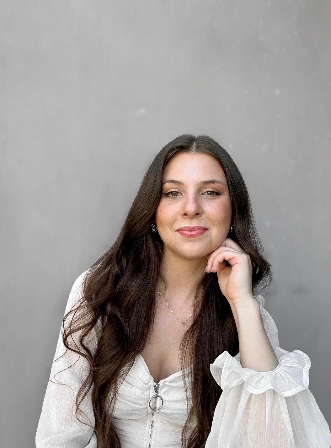 A young woman with long dark hair wearing a white top with a zipper, sitting in front of a plain grey background, smiling softly with her hand resting on her chin.