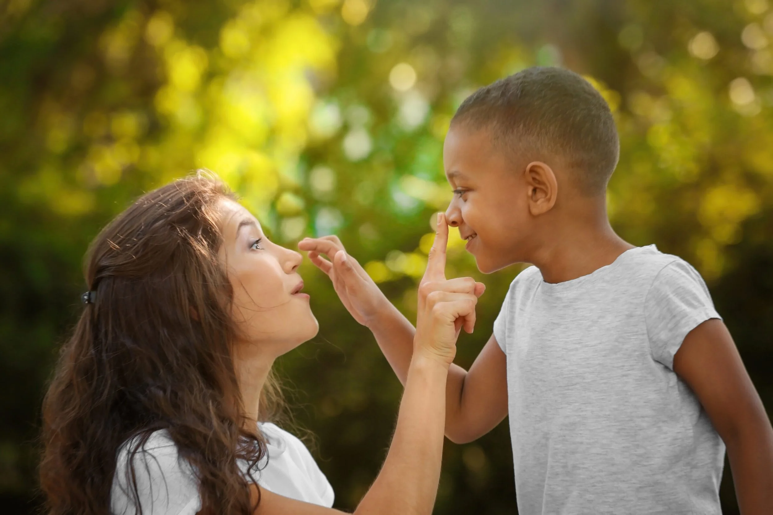 A woman and a young girl are outside in a natural setting. The woman is crouching slightly, with curly brown hair, and the girl has short hair and is smiling. The girl is touching her nose with her finger, and the woman is gently touching the girl's nose with her finger, both looking at each other.