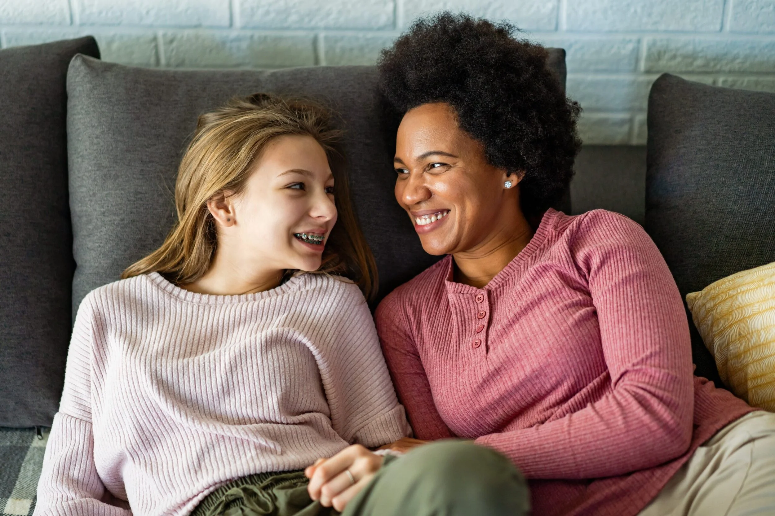 A mother and daughter sitting on a dark gray sofa, smiling and looking at each other, sharing a joyful moment.