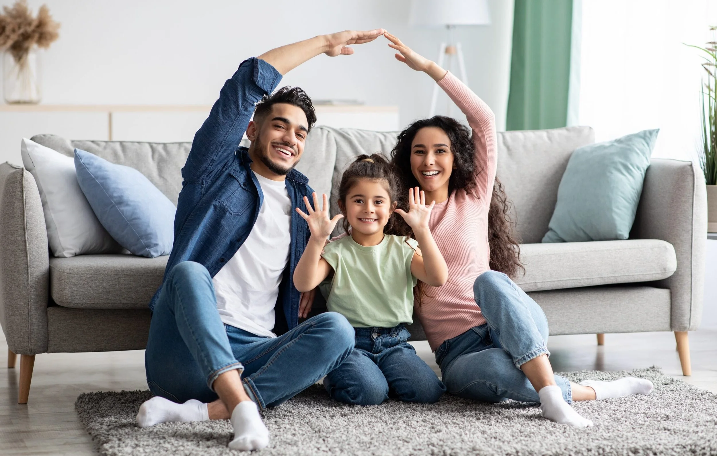 A happy family of three sitting on the floor in a living room, with the parents holding their hands above the child's head to form an arch. They are smiling and waving at the camera.