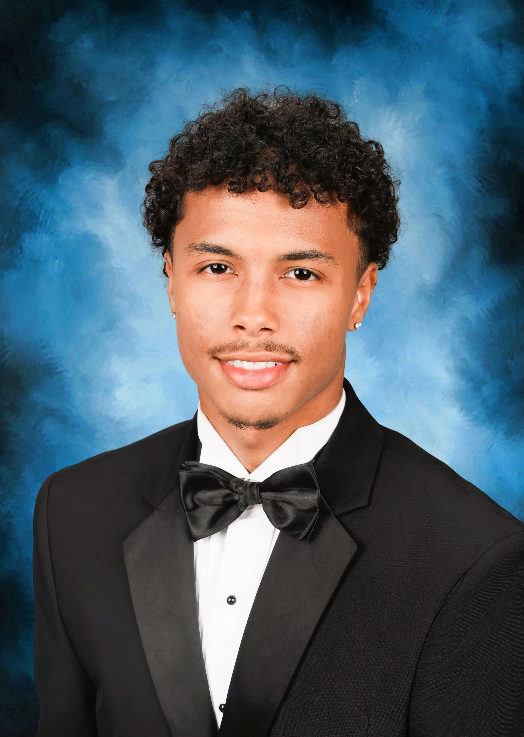A young man in a black tuxedo with a bow tie, smiling, against a blue background.
