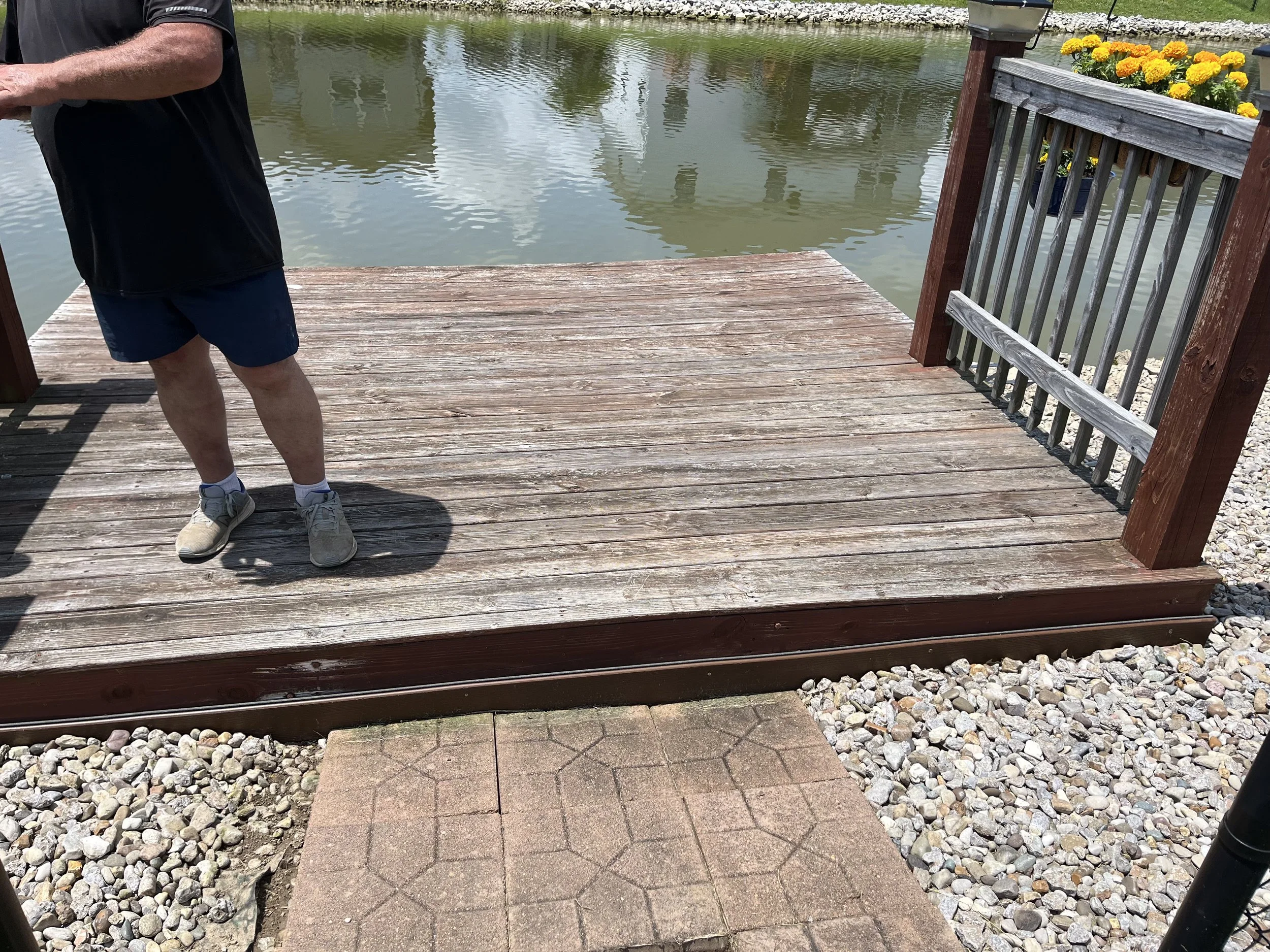 Part of a man wearing black shirt, blue shorts, and white shoes standing on a wooden dock beside a body of water with a stone edge and a flower box with yellow and orange flowers on the railing.
