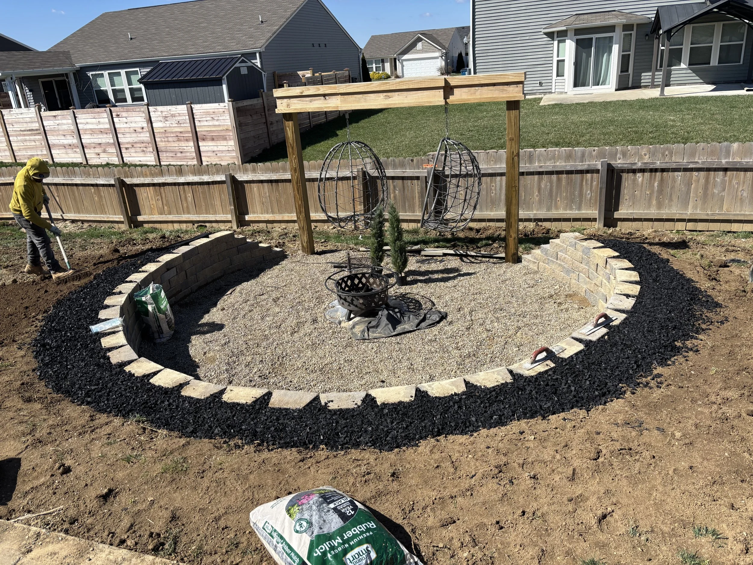 In a backyard, a person wearing a yellow hoodie is working on landscaping. There is a circular garden bed with a stone border, some small plants, and a bag of garden soil. Behind, a wooden structure with hanging spherical wire baskets is visible, and the area is fenced with a wooden privacy fence. Several neighboring houses are visible in the background.