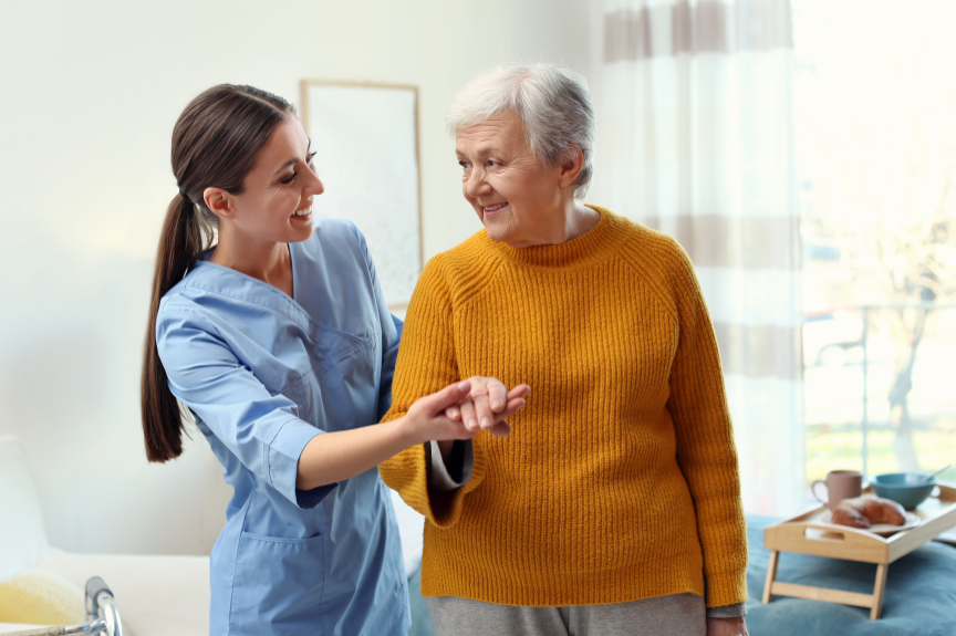 A young female nurse is smiling and helping an elderly woman in her home, both appear happy and engaged in a caring interaction in a bright, cheerful room.