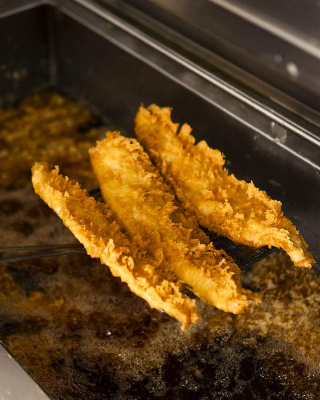 Three pieces of breaded and fried fish being lifted from a fryer or oil with a slotted utensil.
