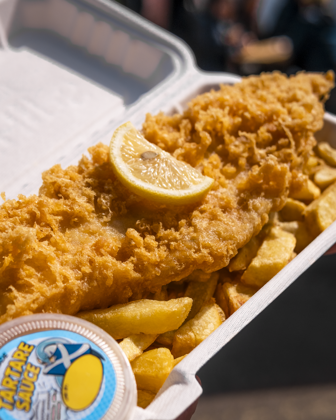 Fried fish fillet with a lemon wedge on top, served with French fries and coleslaw in a takeout container.