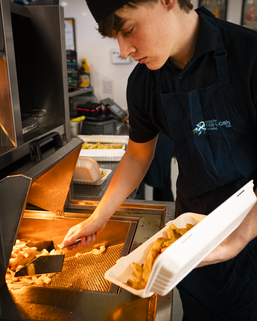 A young man in a black uniform and apron, working in a fast-food restaurant, placing fried food into a white takeout container.
