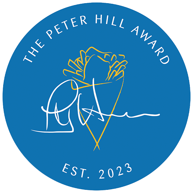 A circular blue logo for The Peter Hill Award, established in 2023, with a stylized gold and white illustration of a mountain and a signature across it.