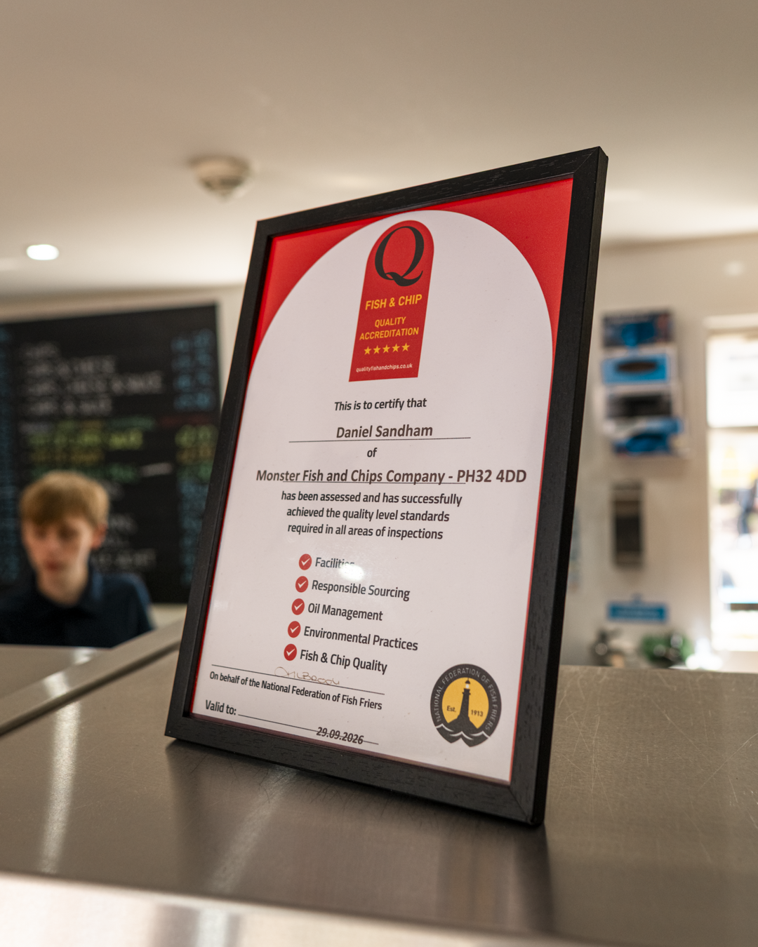 A framed certification plaque on a counter at a fish and chips shop, certifying quality standards, with a young boy blurred in the background.