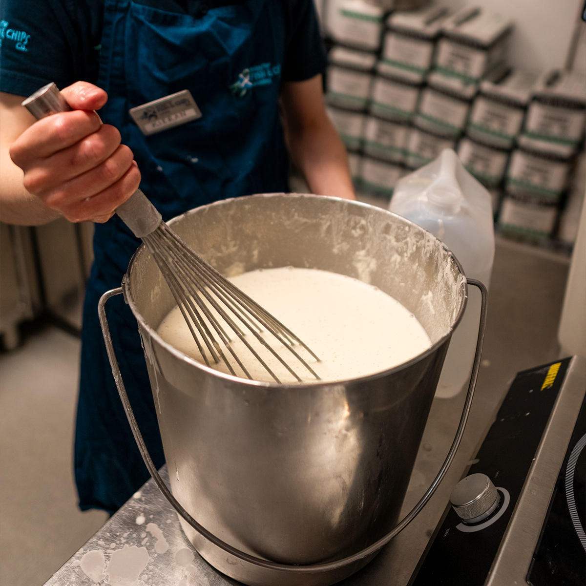 Person stirring a large metal pot of creamy liquid with a whisk in a kitchen or industrial setting.