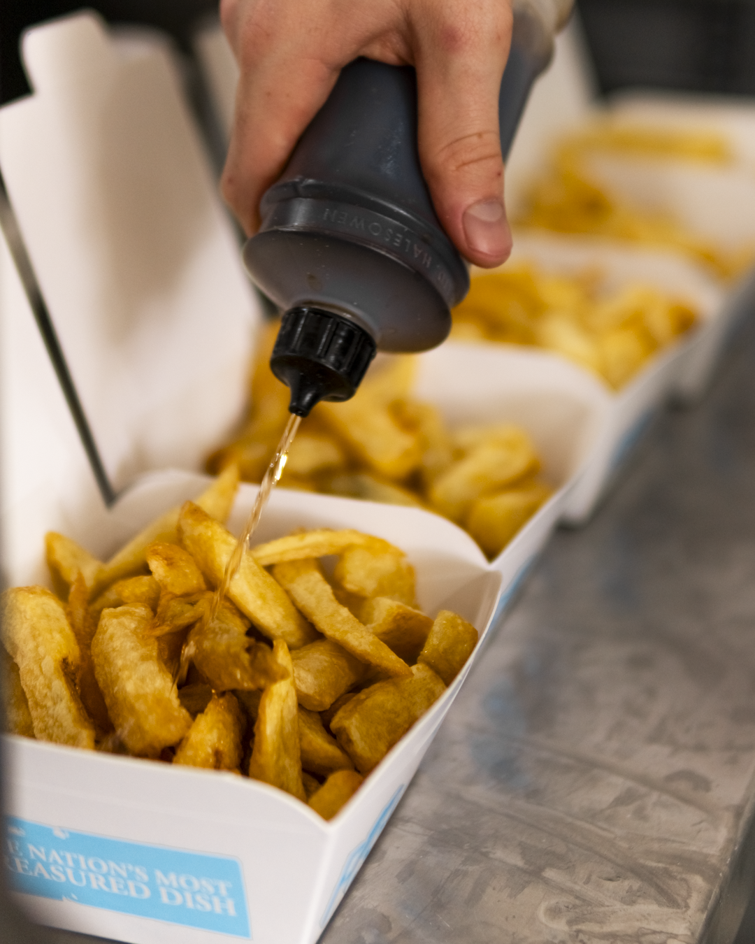 A hand pouring ketchup onto a box of crinkle-cut French fries.