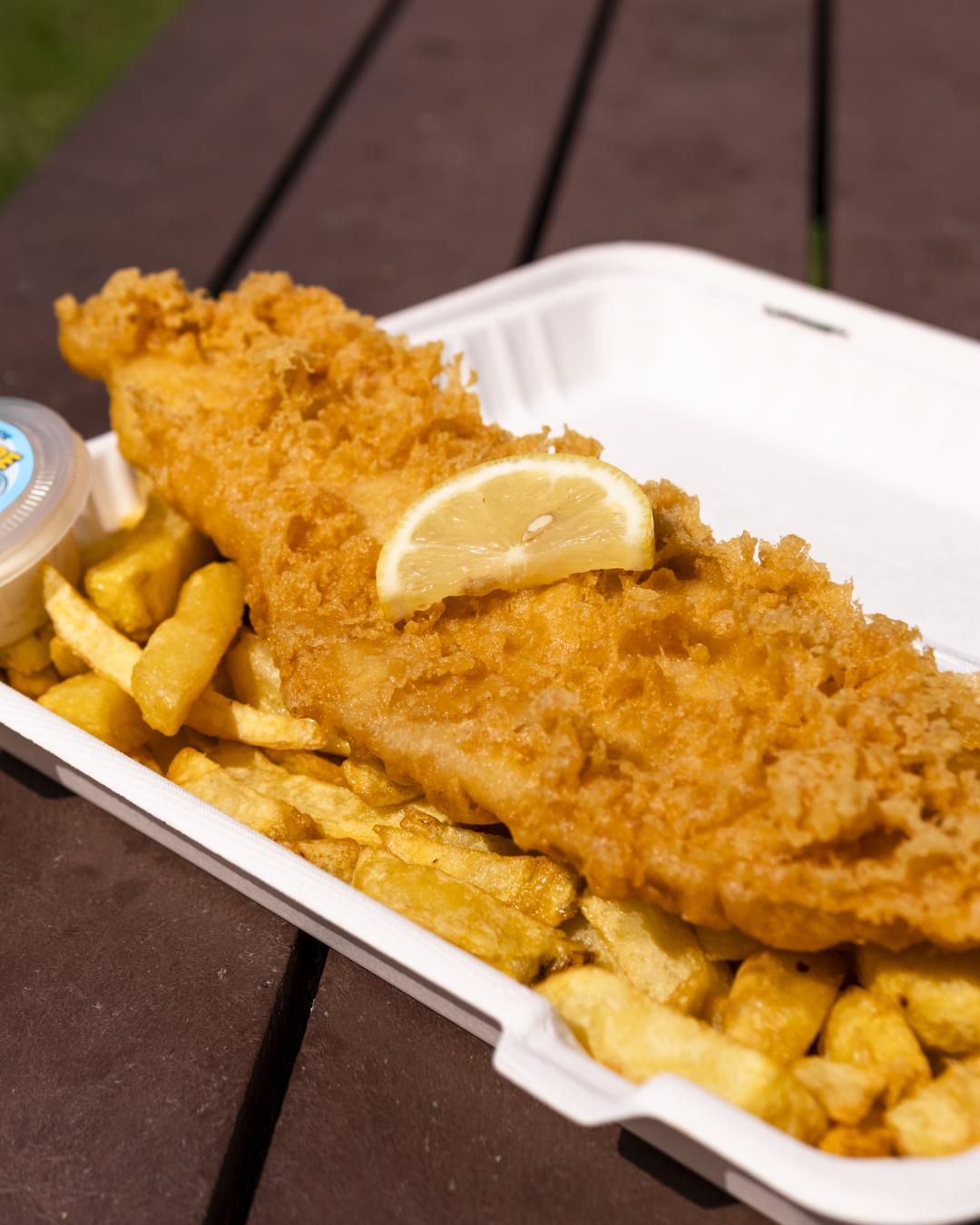Fried fish fillet with a lemon wedge on top, served with French fries, in a takeout container.