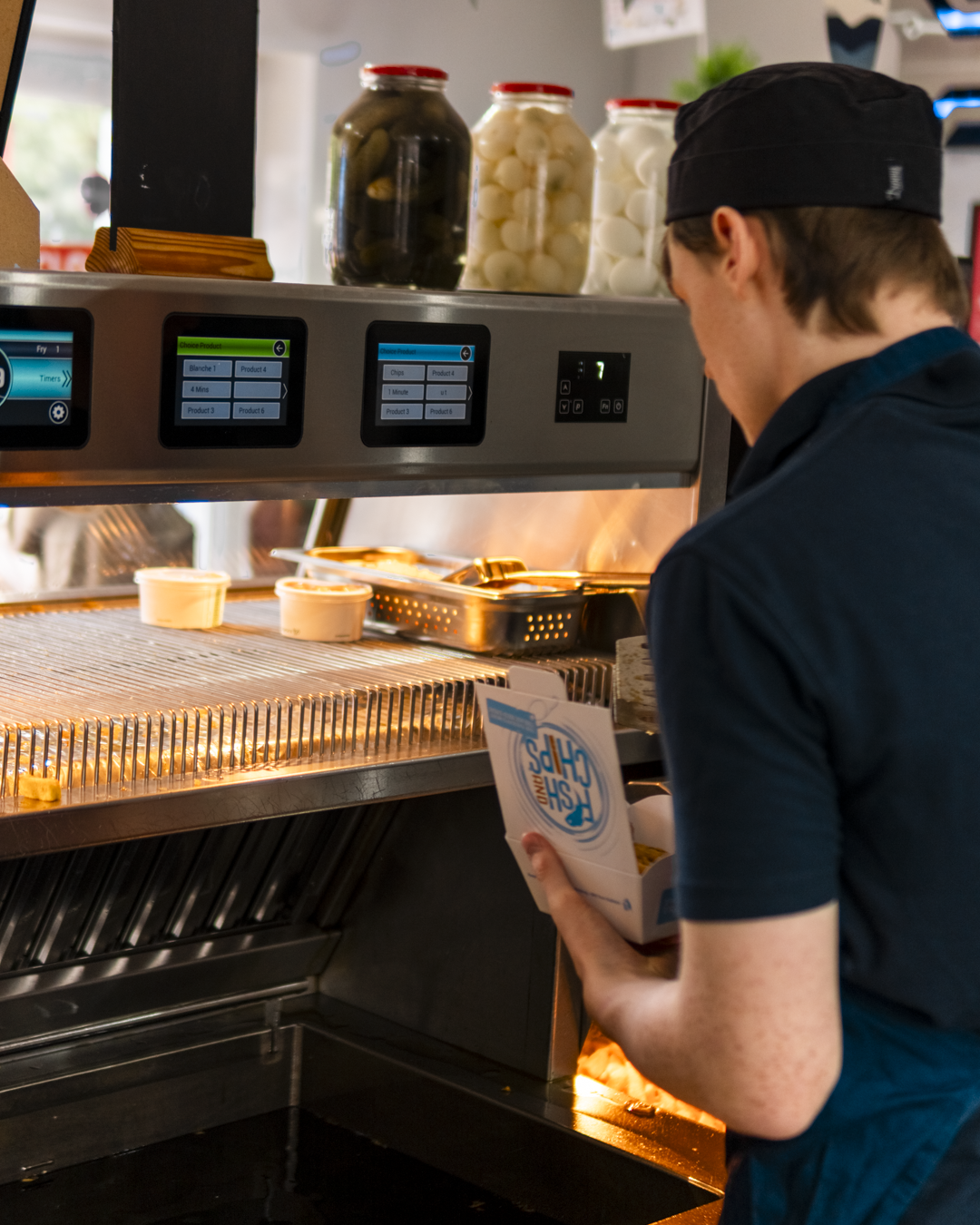 A worker wearing a black uniform and cap loading food containers into a microwave oven at a fast-food restaurant.