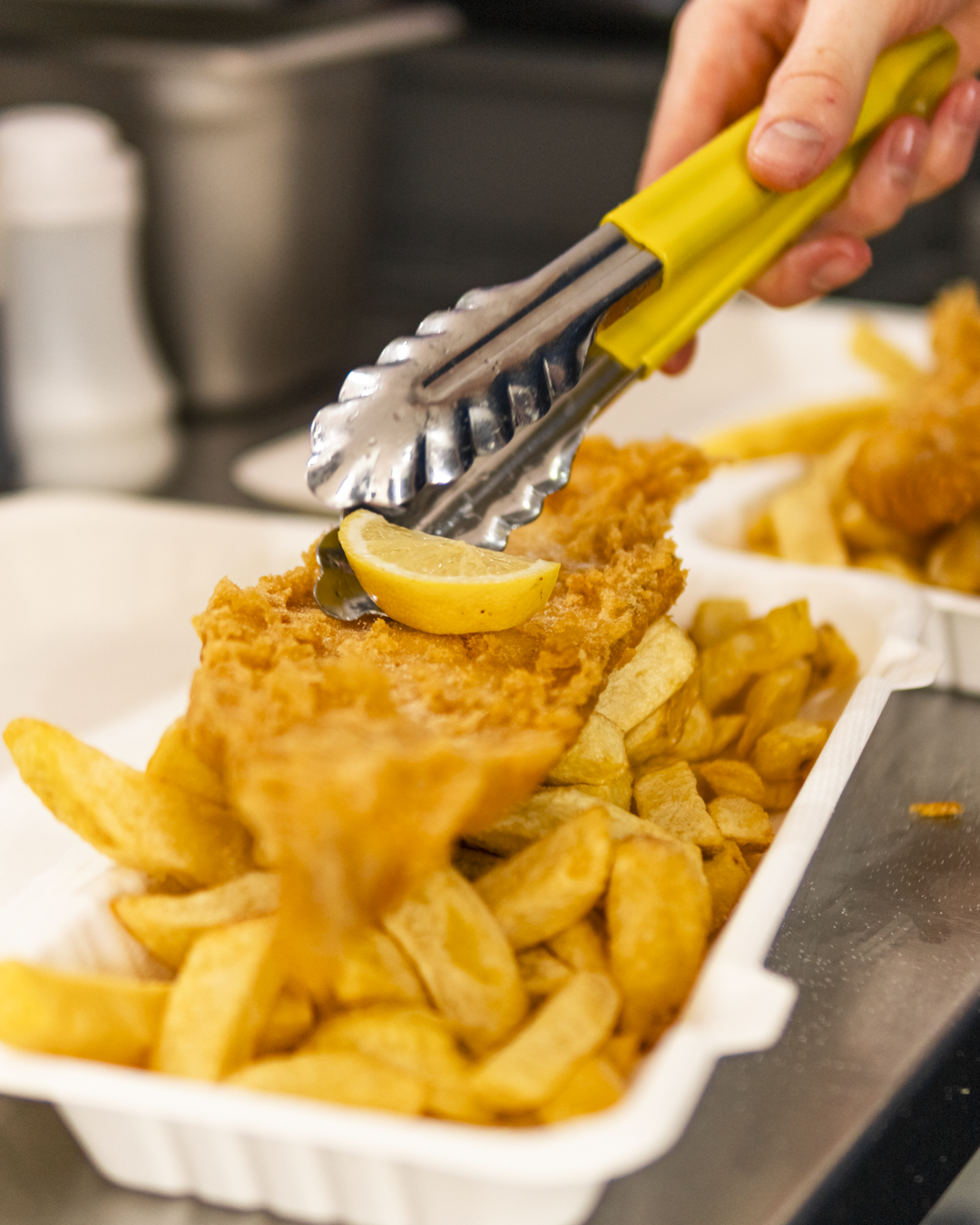 A person using tongs to add a lemon wedge onto a piece of fried fish served with fries in a white tray.