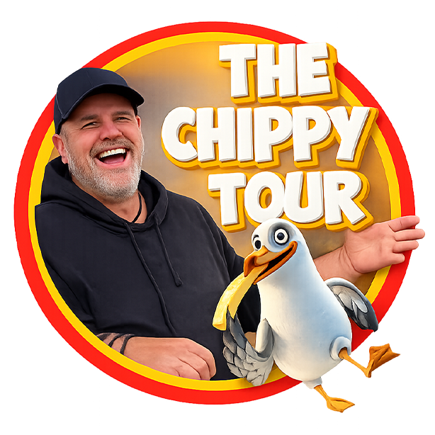 A smiling man with a beard and wearing a black hoodie and black cap, posing with a cartoonish seagull character in front of a sign that says "The Chippy Tour."