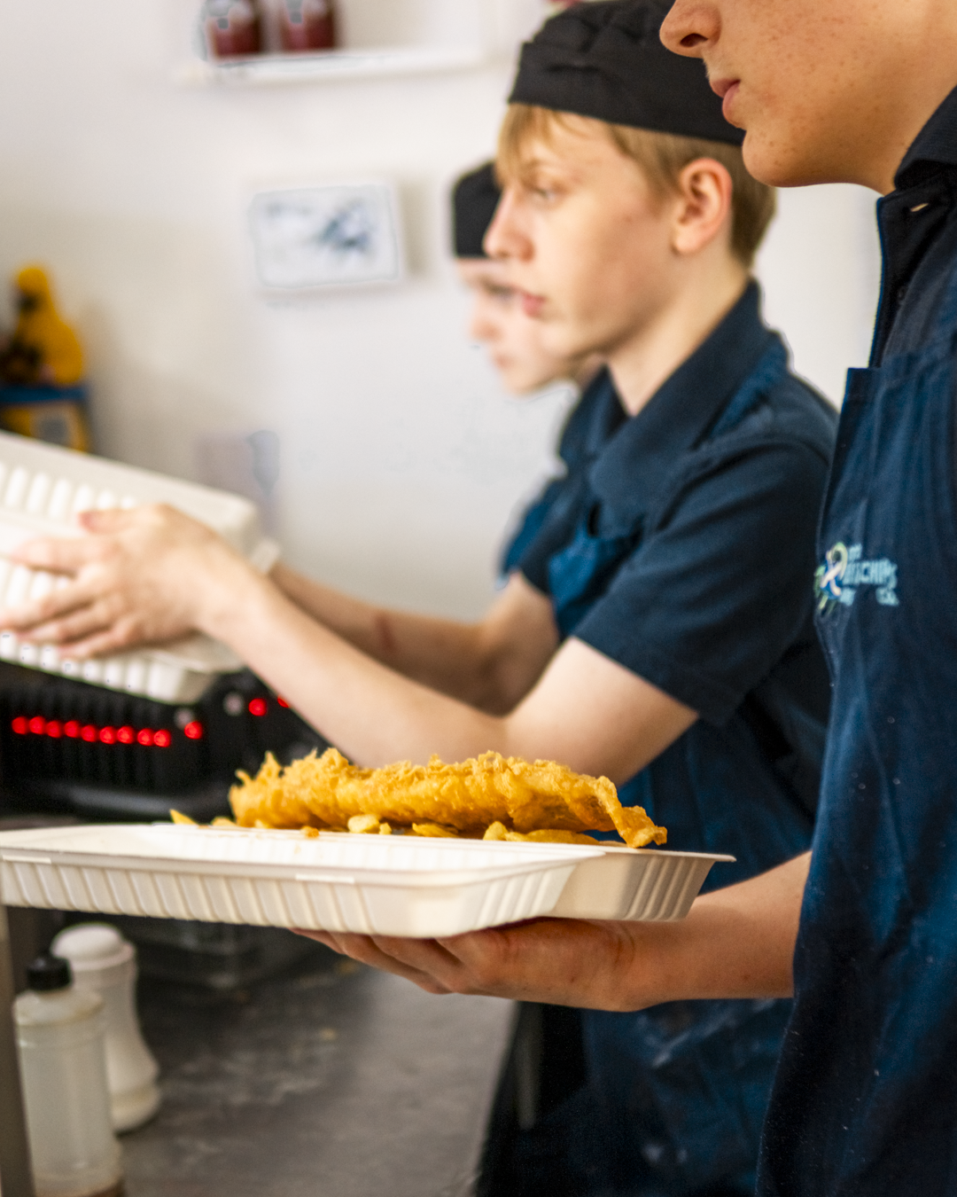 Person holding a foam tray with fried food, with other individuals in the background working in a kitchen or food service setting.