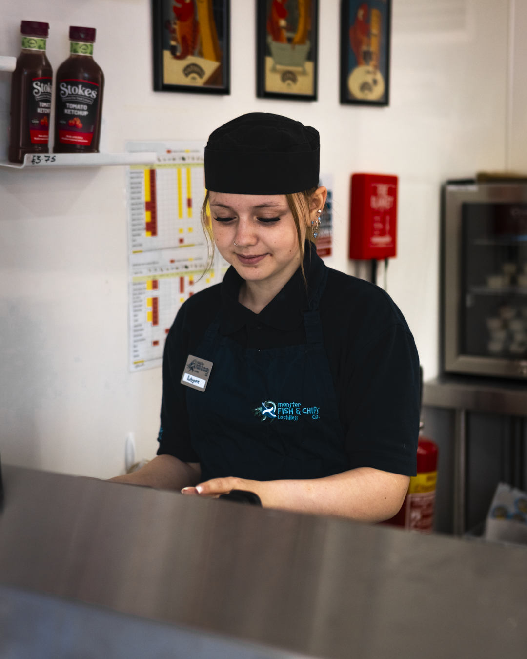 Young woman working behind the counter at a fish and chips shop, wearing a black uniform and apron, with framed artwork on the wall behind her.